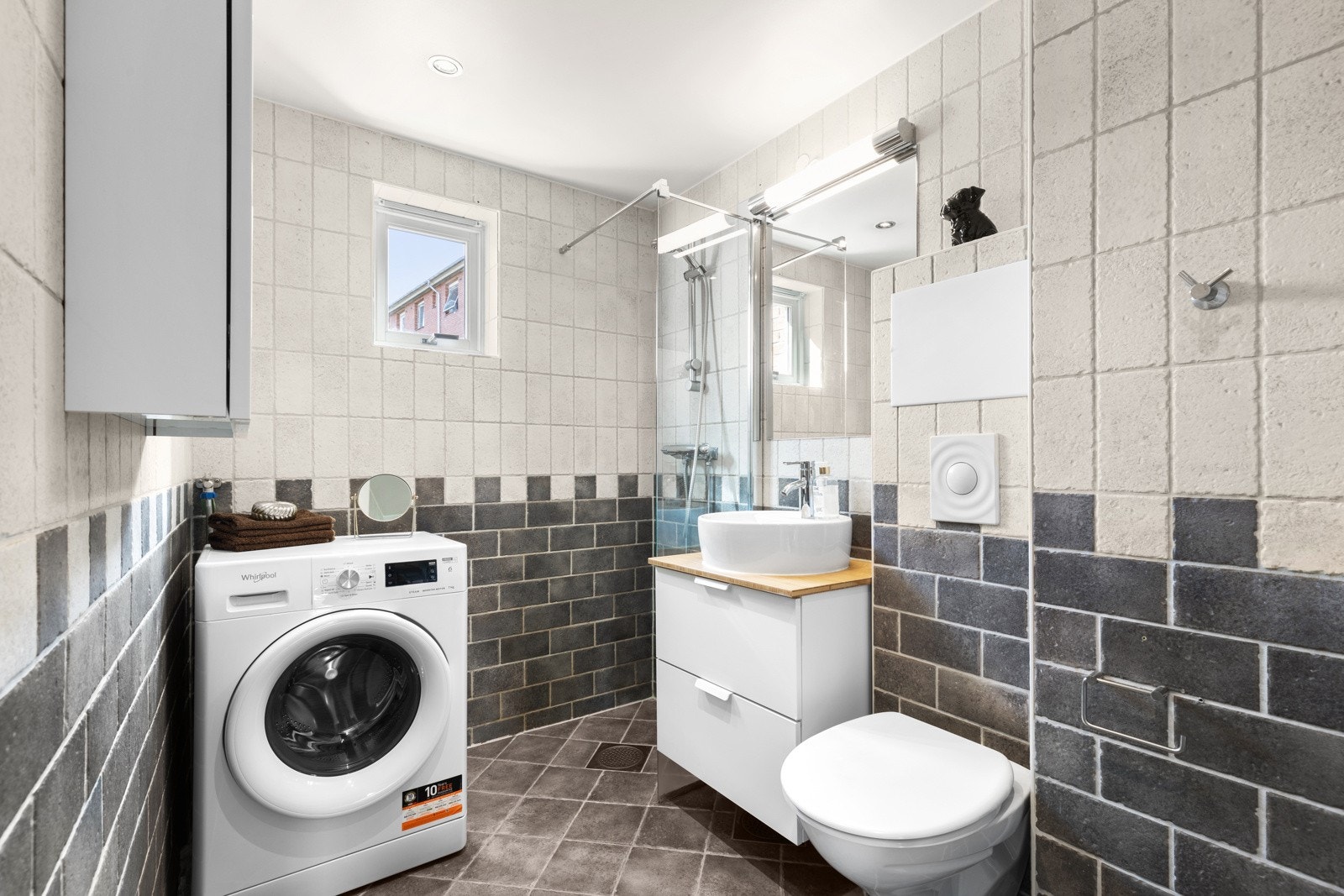 Innredningen består av en toppmontert servant på en skuffeseksjon, et veggskap og et stort speil med belysning. Videre har badet et vegghengt toalett, et dusjhjørne og opplegg for vaskemaskin. Galleribilde