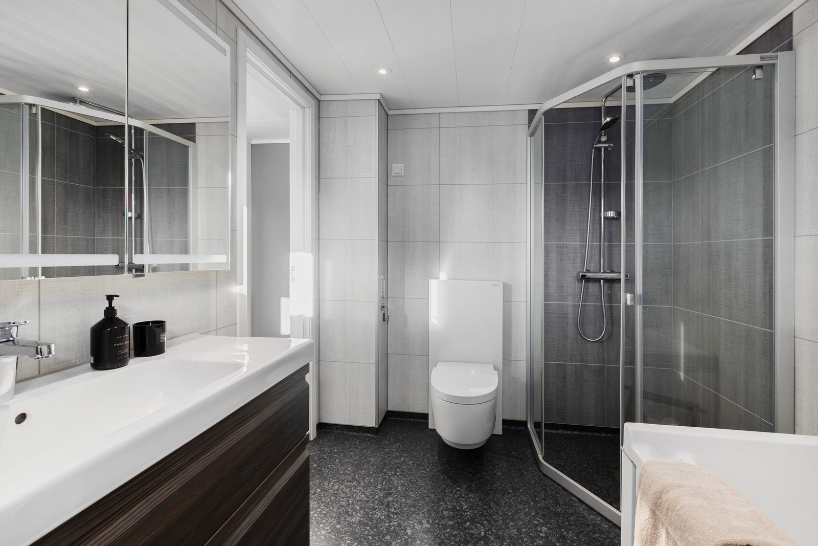 Badet er utstyrt med dusjhjørne, veggmontert spyletoalett, badekar, servant, servantskap og mekanisk avtrekk. Galleribilde