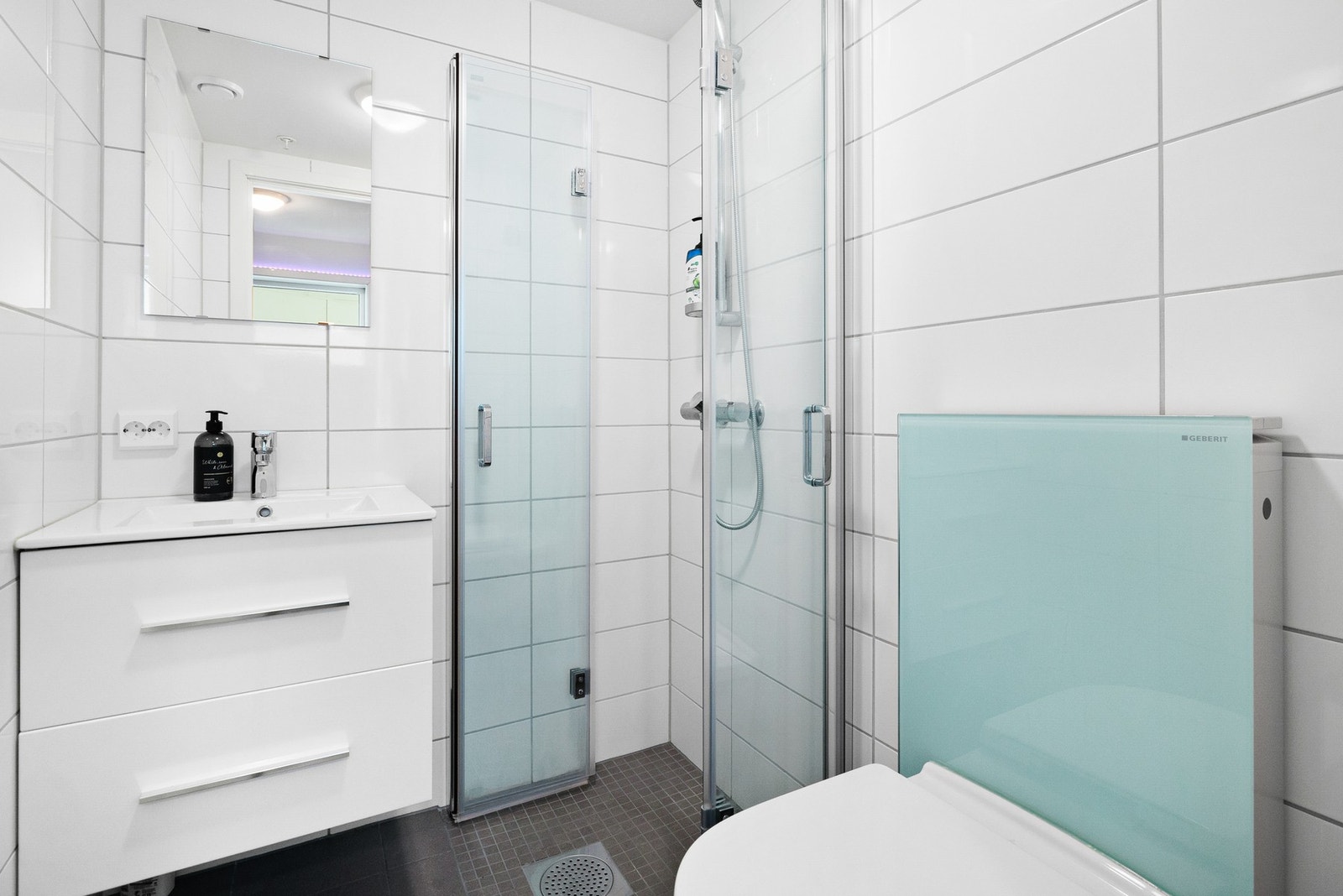 Bad i hybel med flislagte gulv og vegger - utstyrt med dusjhjørne, vegghengt toalett og baderomsinnredning Galleribilde