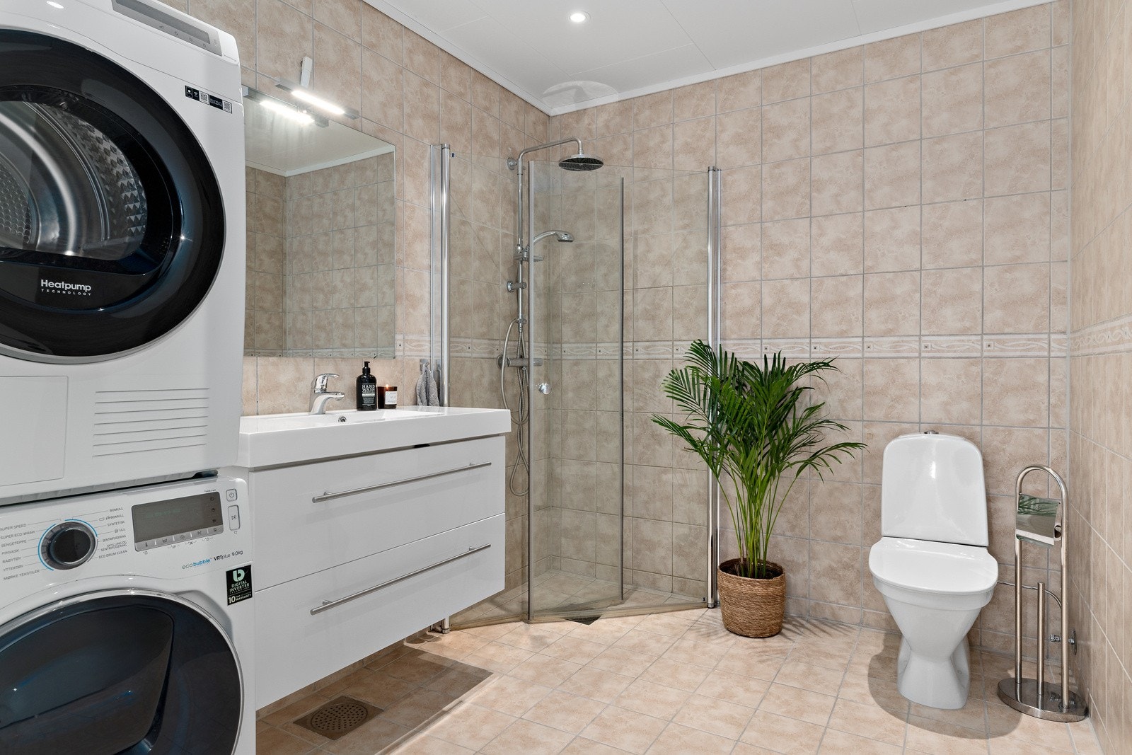 Flislagt bad utstyrt med dusjhjørne, gulvstående toalett og servant, supplert med et romslig servantskap. Galleribilde