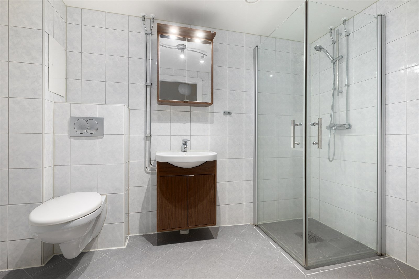 Pent flislagt bad fra 2014 med varmekabler og innredning bestående av vegghengt wc, dusjhjørne innrammet i glassvegger med dør, servantskap og speilskap med integrerte downlights Galleribilde