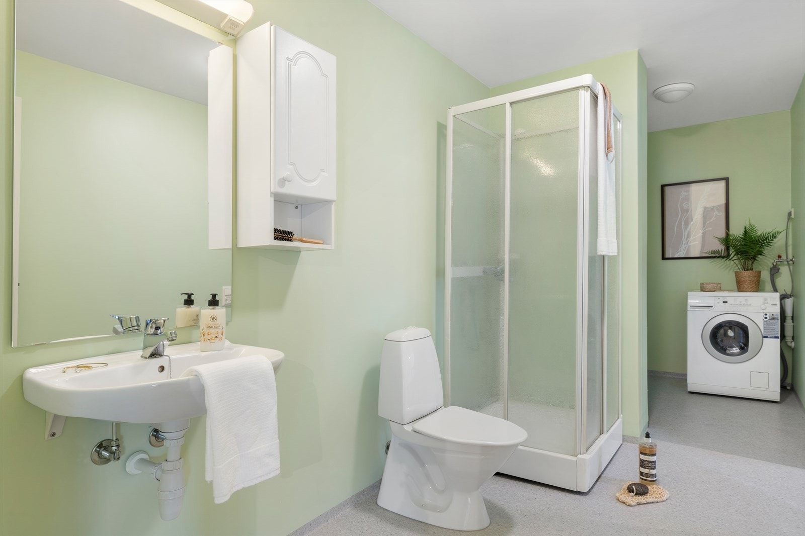 Badet er innredet med toalett, dusjkabinett, vask med speil og belysning over, skap på vegg og fordeler skap for vannrør. Galleribilde