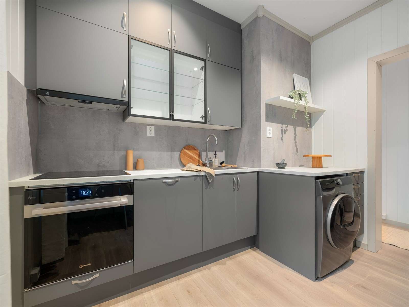 Grå, glattlakkerte fronter samt to Vitrine fronter fra Nettoline kjøkken med laminat benkeplate og nedfelt oppvaskkum. Integrert komfyr med induksjon platetopp, mikrobølgeovn, oppvaskmaskin, vaskemaskin og kjøl/frys. Galleribilde