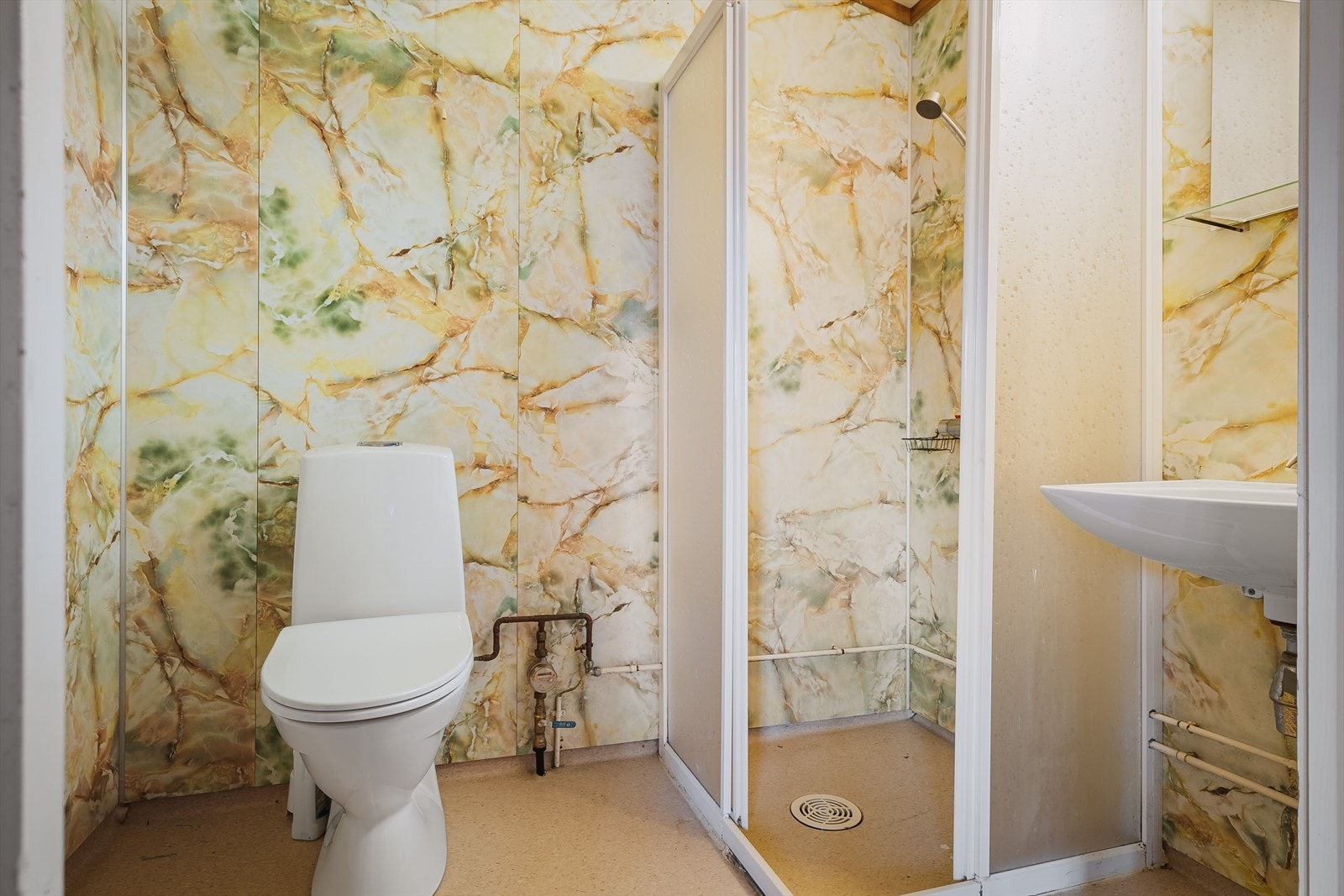 Eldre baderom med belegg på gulv og våtromsplater på veggene. Baderommet er utstryt med vegghengt servant, speil med belysning over, dusjhjørne og gulvstående toalett. Galleribilde