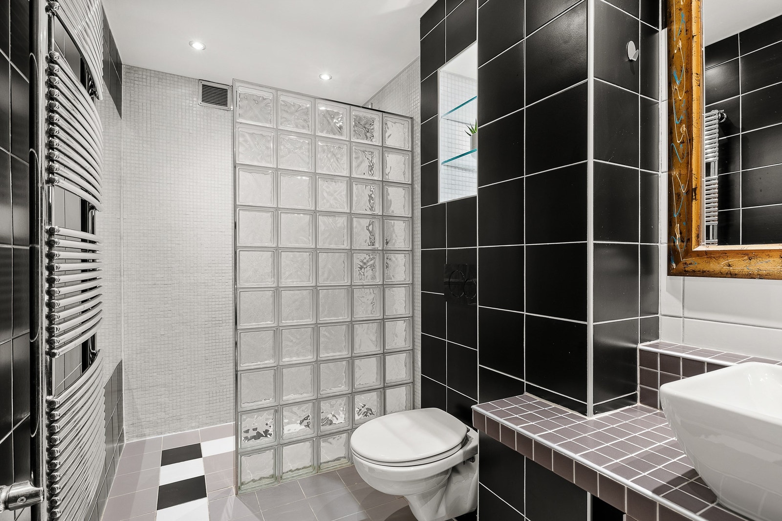 Baderommet er utrustet med flislagt benk med ovenpåliggende servant. Speil på vegg over servant. Dusj med skillevegg av glassbyggersten. Vegghengt toalett med innebygd sisterne. Vegghengt håndkletørker tilkoblet vannbåren varme. Galleribilde