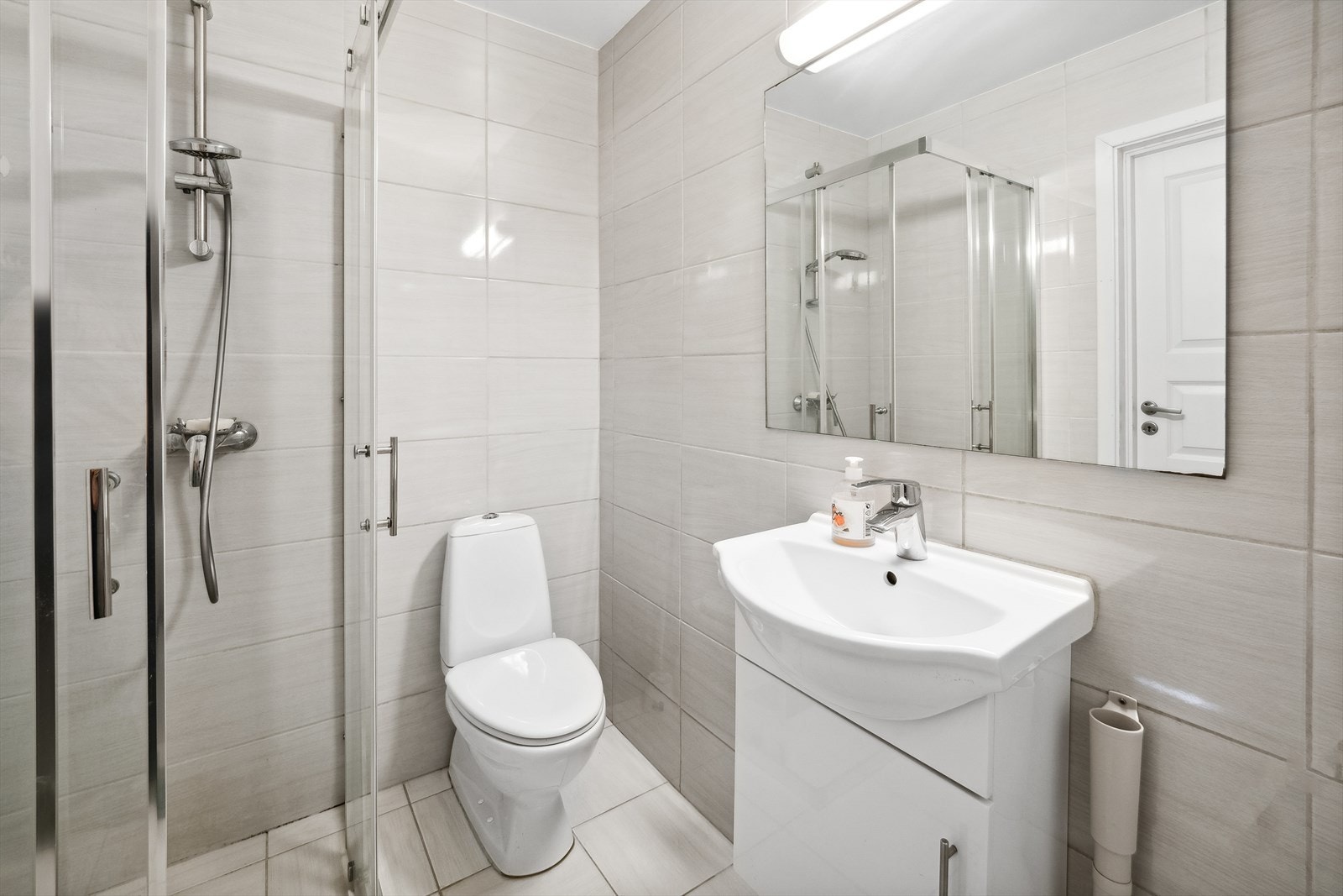 Flislagt baderom med elektriske varmekabler i gulv og malt betong i himling. Badet er utstyrt med servant med underskap, speil med tilhørende belysning, dusjdører og dusjhjørne. Galleribilde
