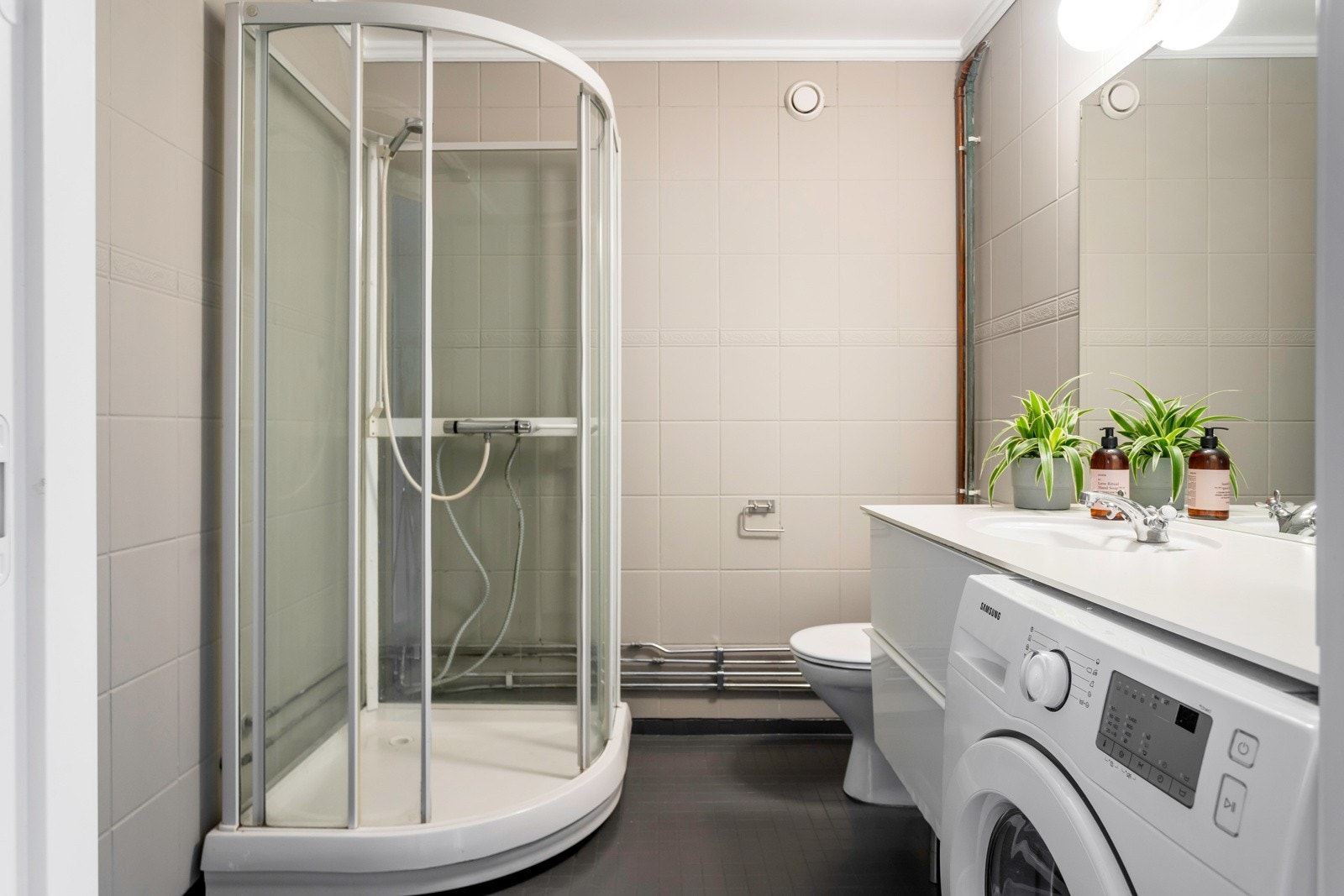Baderom med gulv- og veggflater av malte fliser. Badet er innredet med et dusjkabinett, gulvstående toalett, og servantskap. Det er opplegg for vaskemaskin på badet. Galleribilde