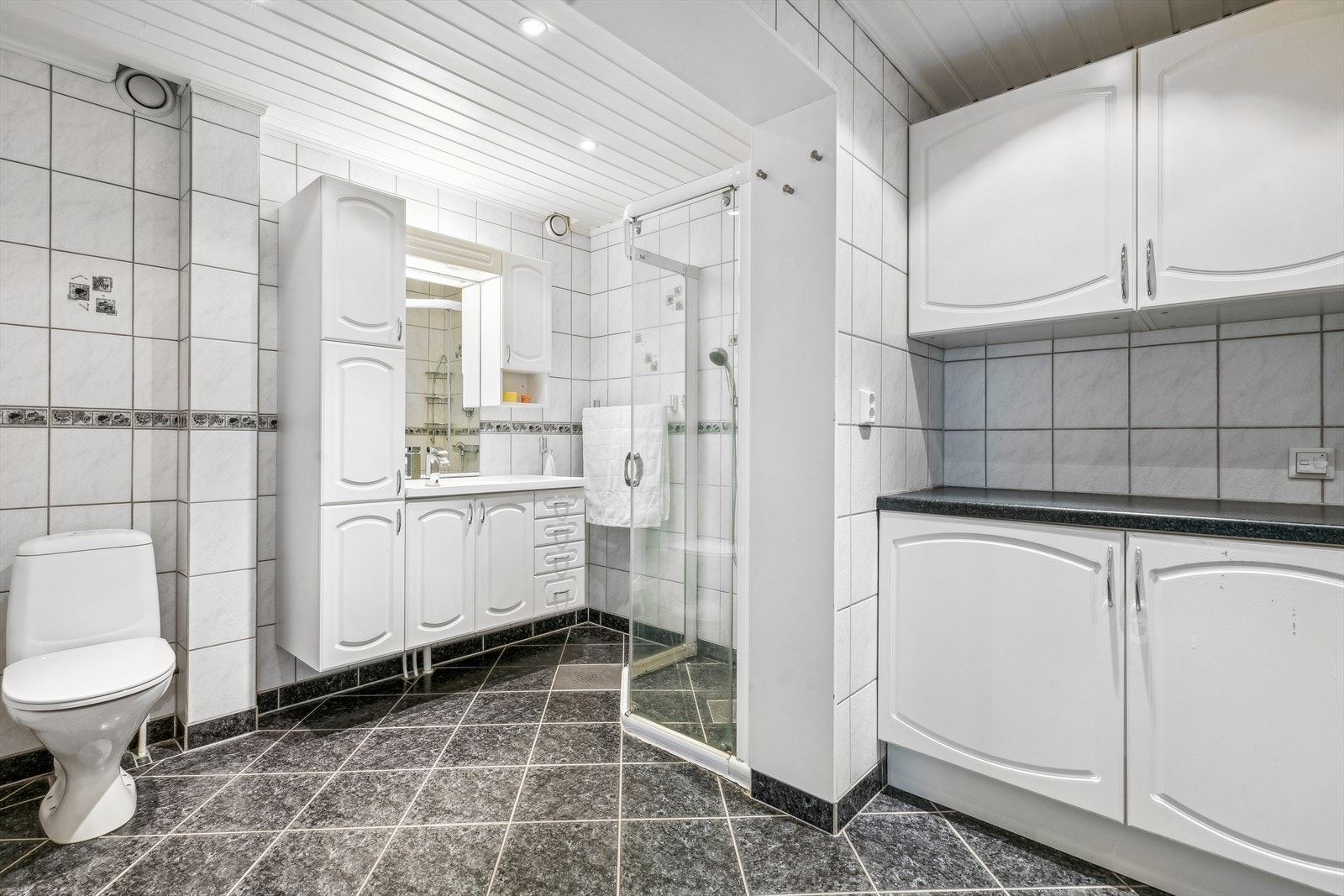 Her er det innredet med gulvmontert toalett, dusjhjørne, samt en baderomsinnredning med hvite profilerte fronter og heldekkende servant. Galleribilde