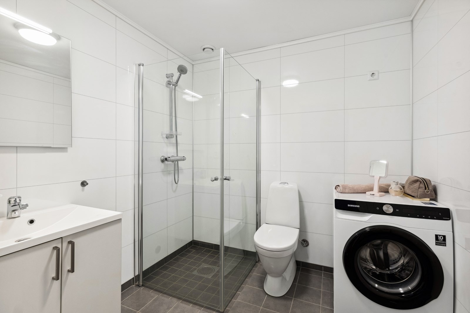 Badet er utstyrt med dusjhjørne med innfellbare glassvegger, gulvstående wc, servantinnredning med underskap, samt opplegg og plass til både vaskemaskin og tørketrommel. Galleribilde