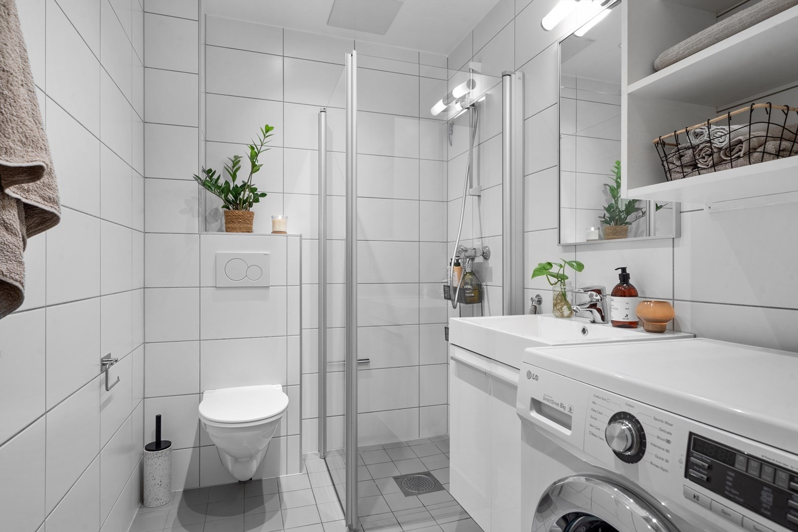 Pent bad med lyse fliser, hjørnedusj med glassdører, vegghengt toalett og servant med oppbevaring. Galleribilde