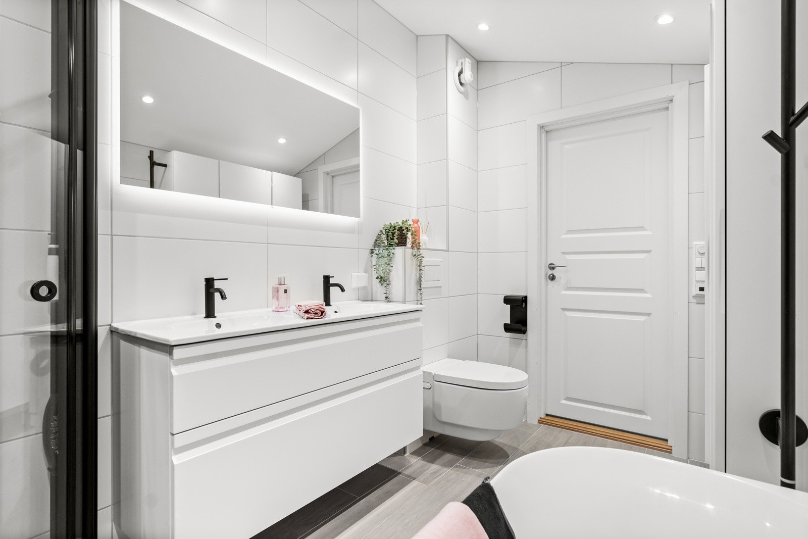 Badet inneholder servantinnredning med skuffer og dobbel servant, speil med belysning, badekar, vegghengt wc og dusjhjørne. Galleribilde