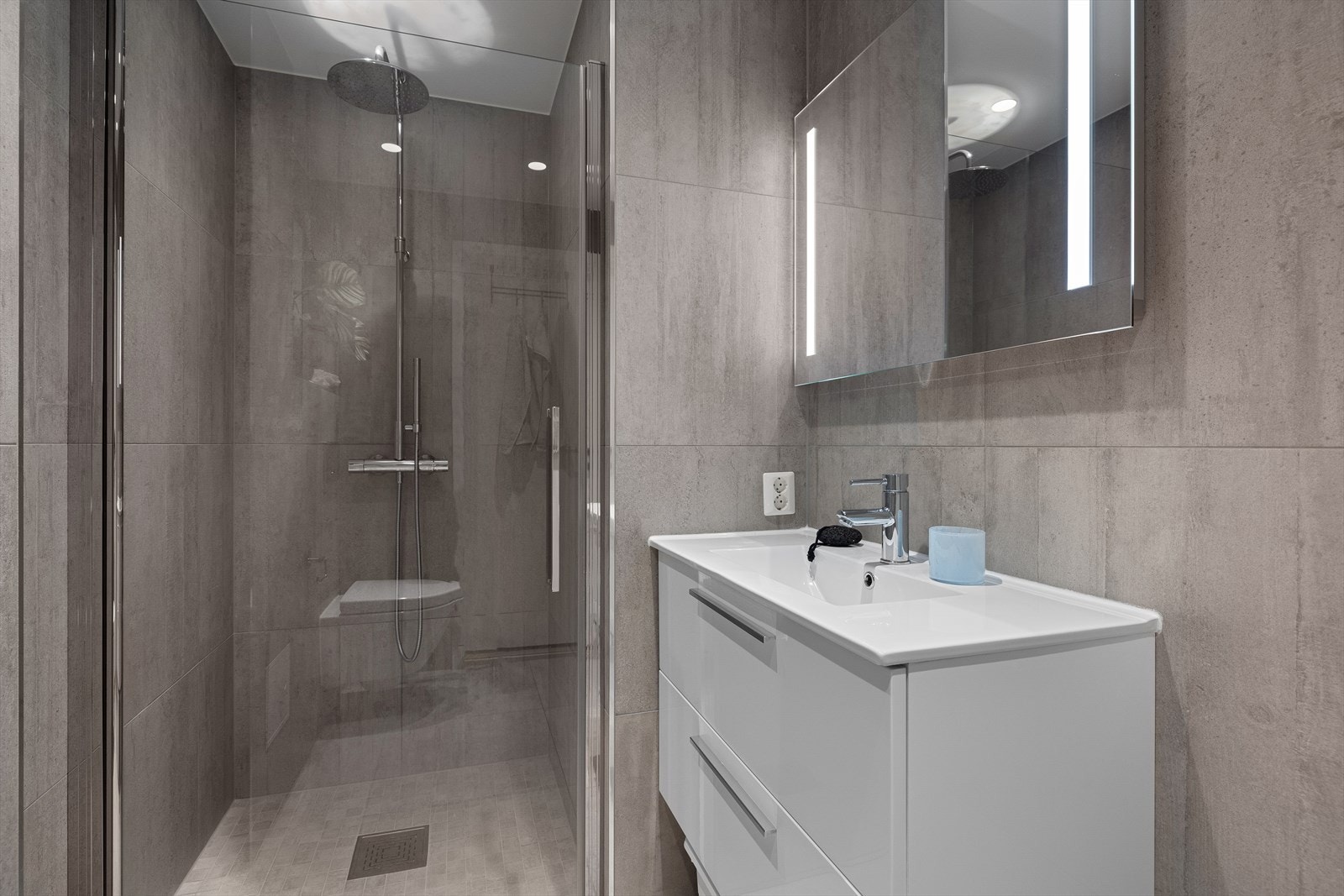 Badet er utstyrt med dusjhjørne med glassdør, veggmontert toalett og baderomsinnredning med skuffer, overmontert servant, blandebatteri fra Tapwell og veggmontert speil over servant med innfelt lys Galleribilde