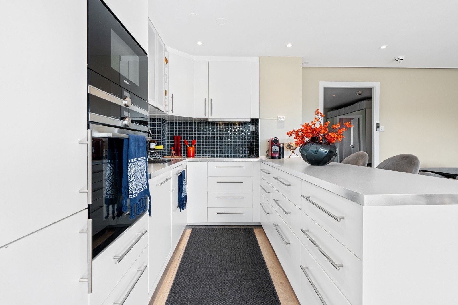 Kjøkkeninnredning går helt opp til taket og består av glatte hvite fronter og vitrineskap. Grå benkeplate, nedfeldt kum i rustfritt stål og mosaikkfliser på vegg. Galleribilde