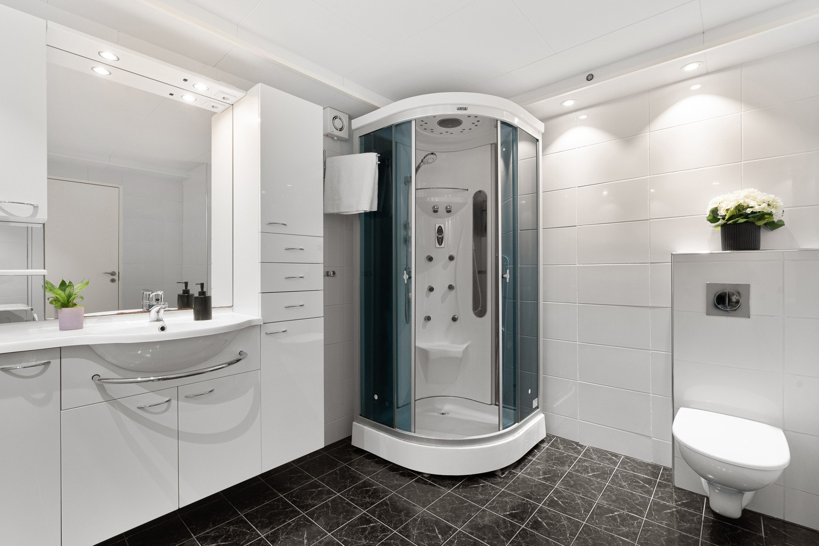 Flislagt bad/vaskerom med gulvvarme, vegghengt wc, lys baderomsinnredning med heldekkende servant og speil med belysning. Galleribilde