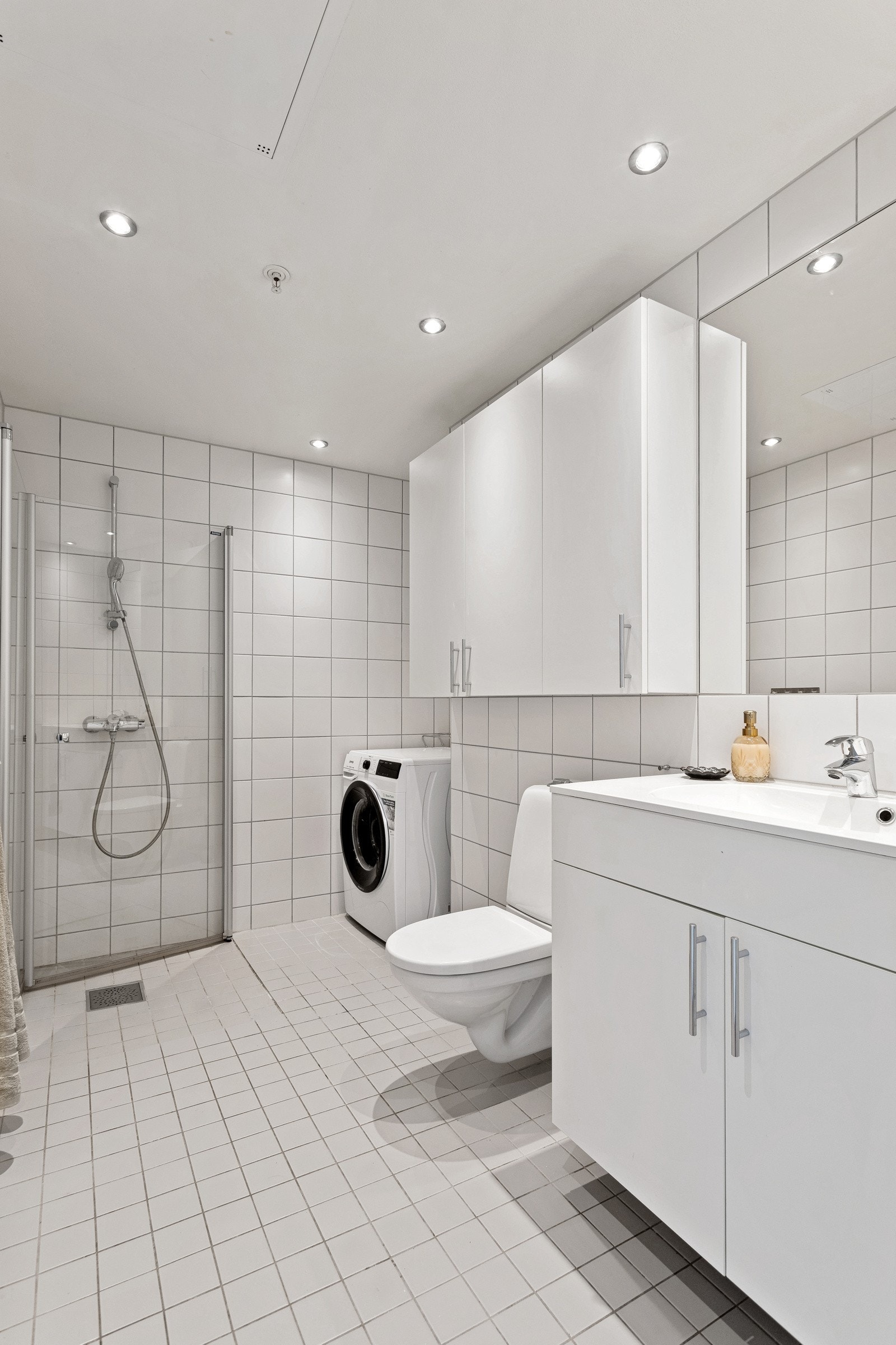 Badet er utstyrt med dusj med inn- utadslående dusjvegger, servant med underskap, speil, veggskap og wc. Opplegg for vaskemaskin. Galleribilde