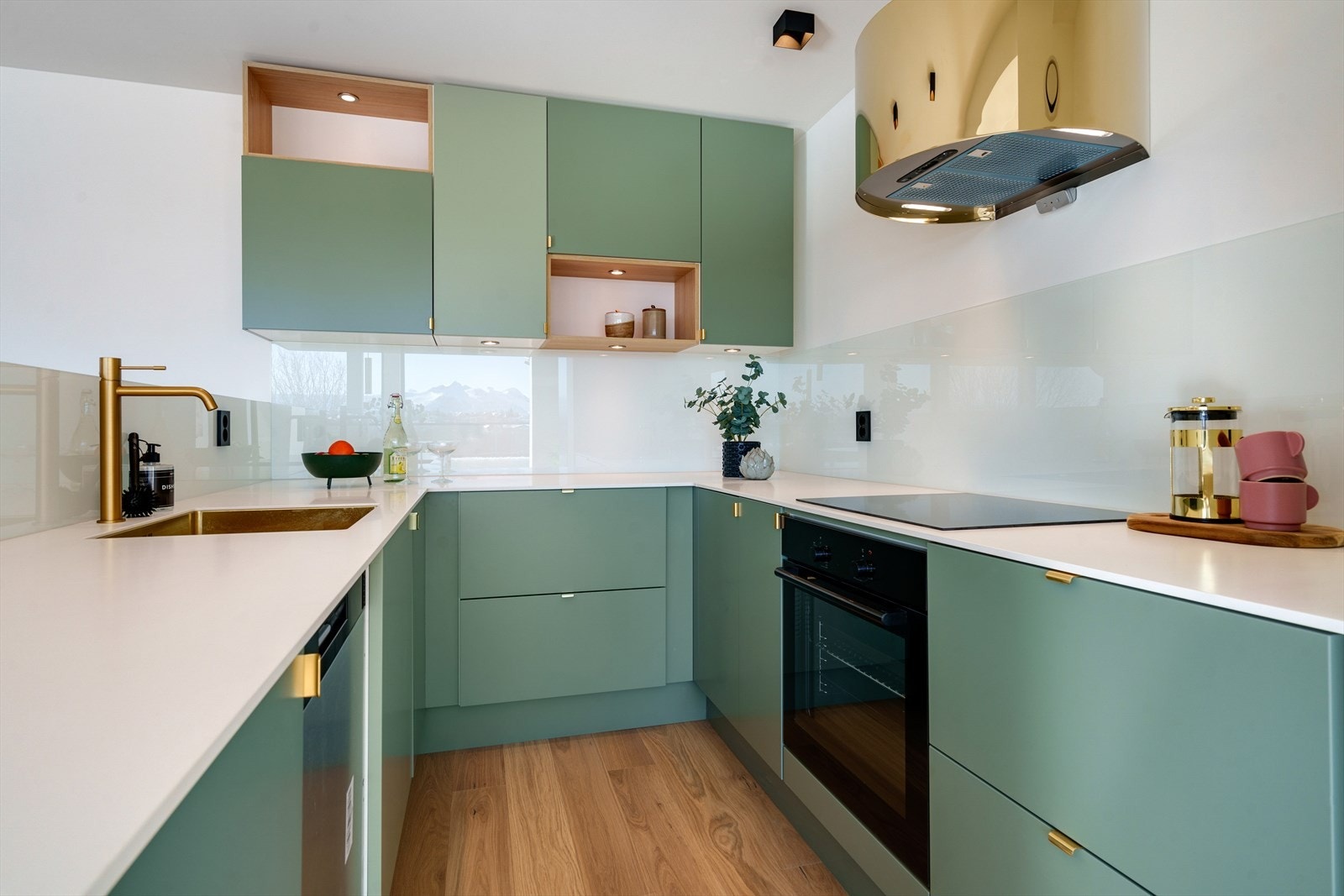 Kjøkkenet har en pen innredning med slette fronter i en moderne og tidsriktig farge m/håndtak i gullfarge - Videre er det hvit benkeplate med underlimt vask og glassplater på vegg over benk Galleribilde