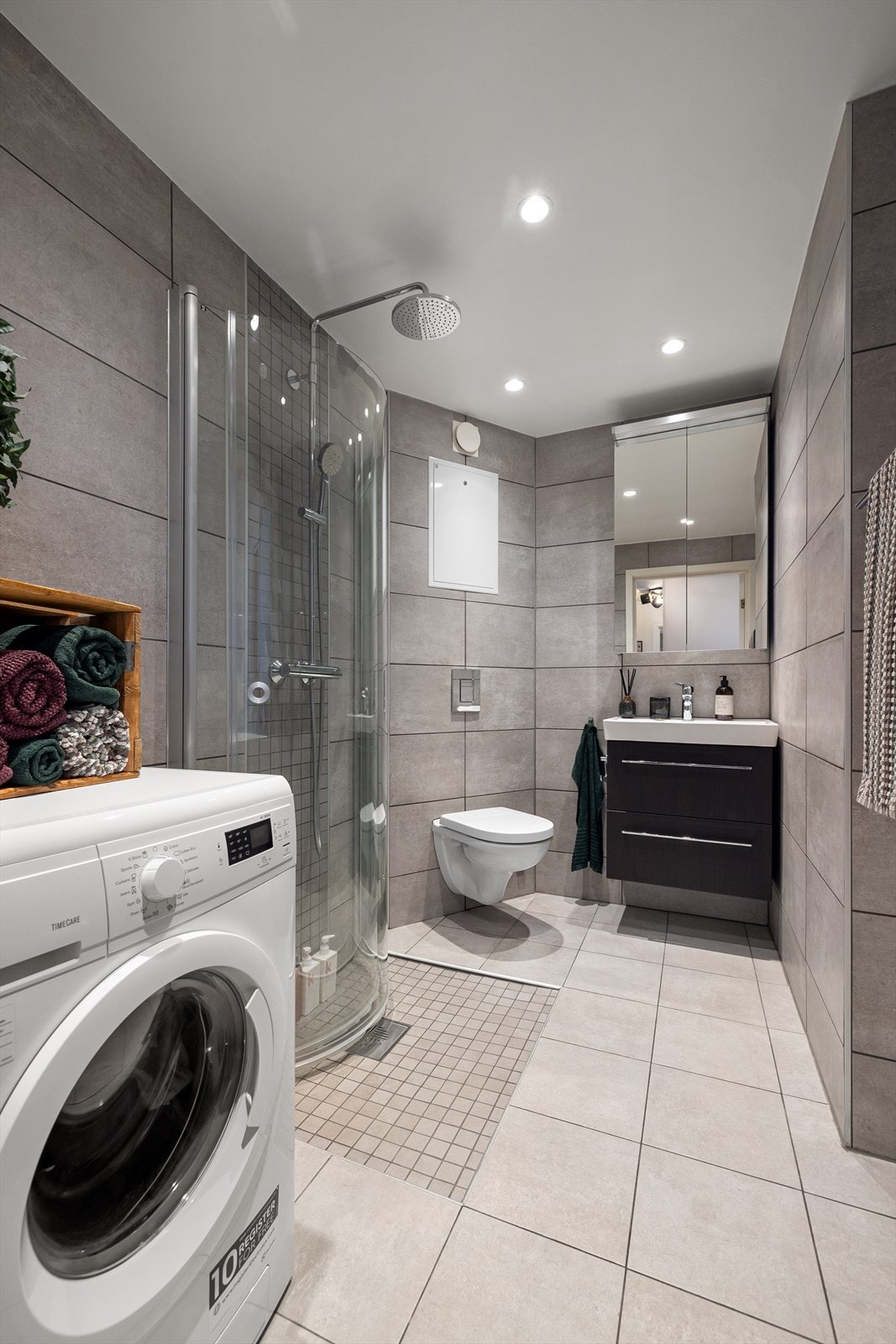 Badet er utstyrt med veggmontert toalett, dusjhjørne med innfellbare glassdører og baderomsinnredning med skuffer, overlimt servant Galleribilde