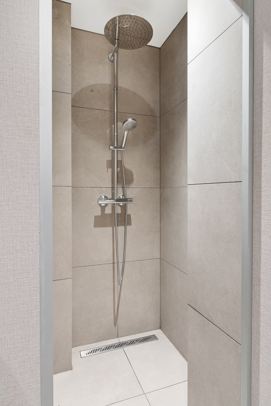 Stor dusjnisje med regnfallsdusj som utnytter romstrukturen godt. Badet har synlig membran under klemring på sluk, en elektrisk vifte i taket og tilluftsåpning under døren som ventilasjonssystem. Galleribilde