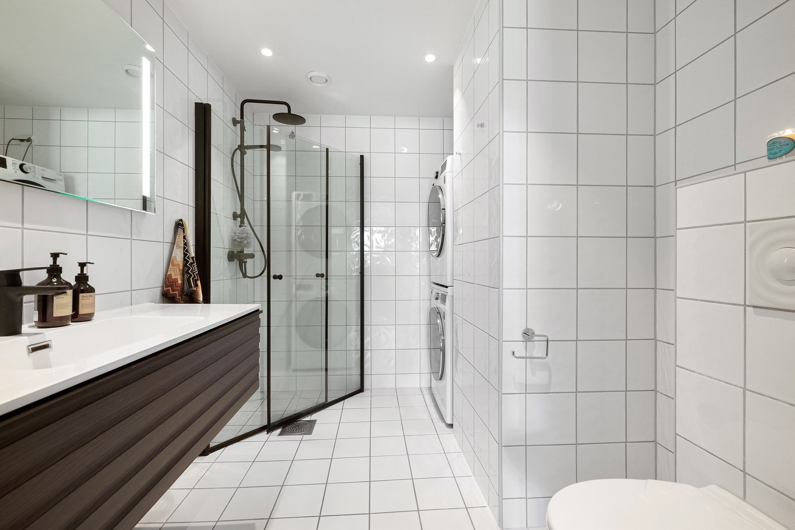 Lekkert og romslig baderom med flislagt gulv med varme, flislagte vegger, og nedsenket himling med malt flate, avtrekksventil og downlights. Galleribilde