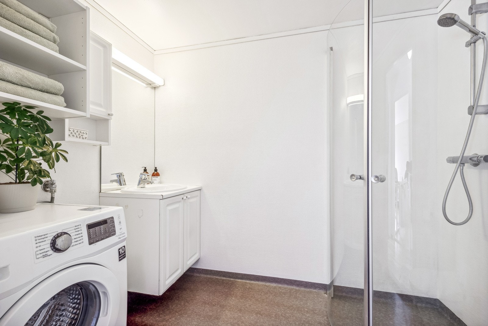 Lyst baderom med varmekabler i gulv og praktiske dusjvegger. Badet har servant i en hvit innredning med speil, belysning samt opplegg for vaskemaskin. Galleribilde