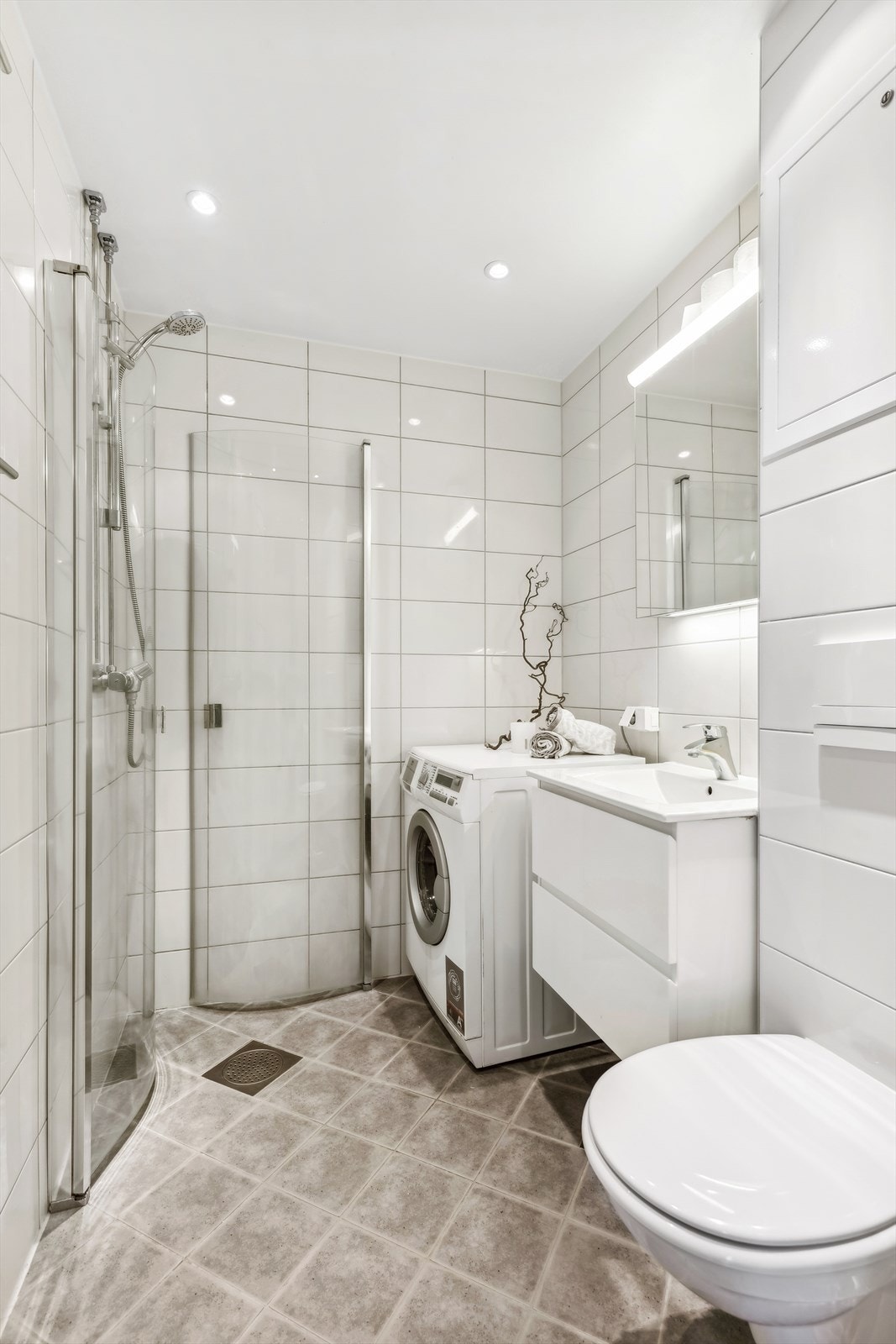 Badet er utstyrt med innadslående dusjdører med diusjsøyle, vegghengt servantseksjon og vegghengt toalett. Avsatt plass til både vaskemaskin og tørketrommel. Galleribilde