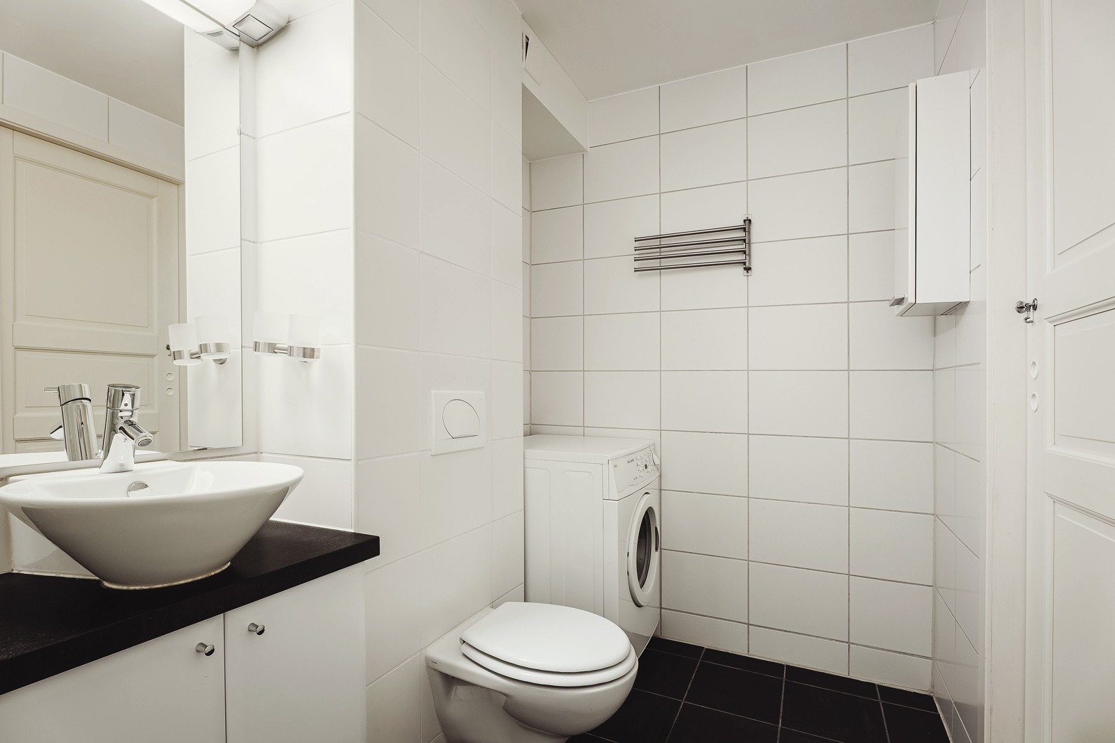 Rommet er utstyrt med veggmontert klosett, toppmontert håndvask med underskap og speil, dusjhjørne med glassdører og opplegg for vaskemaskin og tørketrommel. Galleribilde