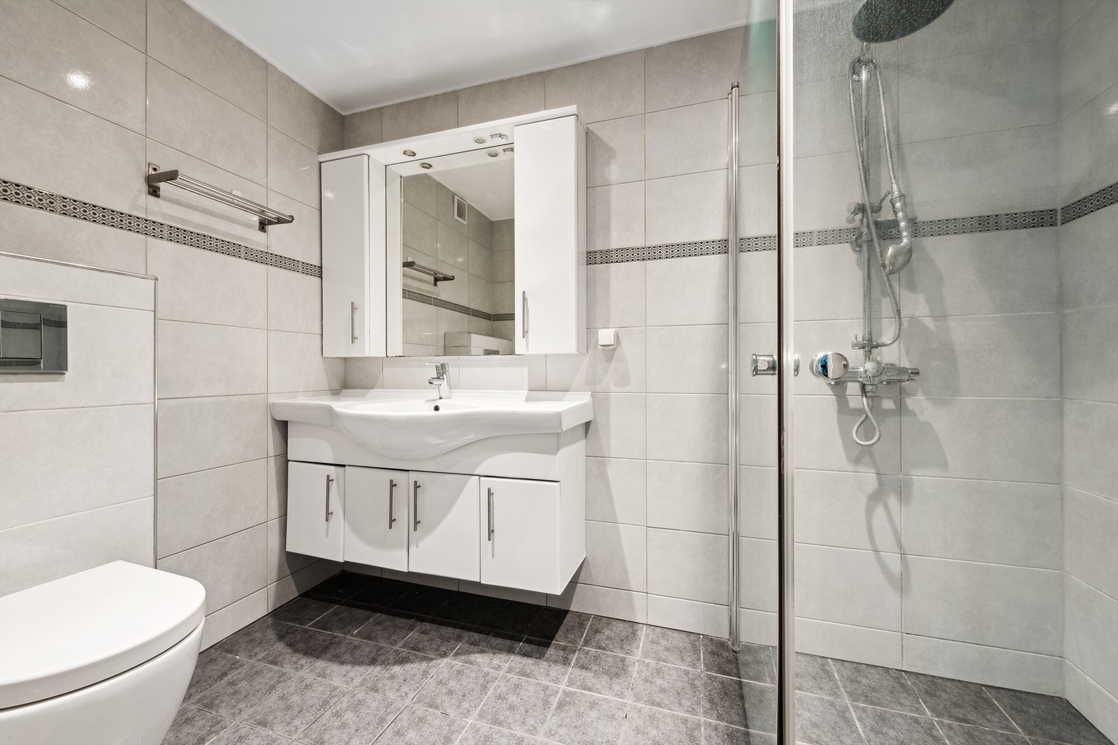 Baderom ved kjøkken fra 2015 med flislagte gulv/vegger og downlights i himling. Badet er utstyrt med servant, dusj, toalett, baderomsinnredning med glatte overflater og speil med lys. Det er plass og opplegg for vaskemaskin. Galleribilde