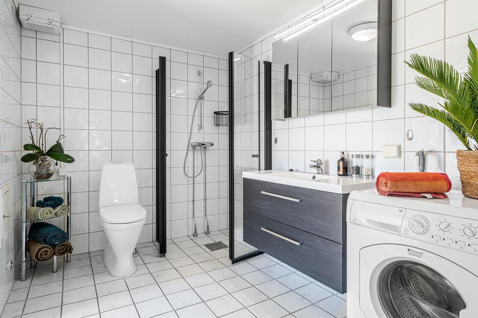 Lyst bad med flis på gulv og vegger. Her er det toalett, dusjhjørne med dusjvegger og innredning med heldekkende hvit vask, underskuffer med slette fronter og overskap med speildører. Badet har også opplegg for vaskemaskin. Galleribilde