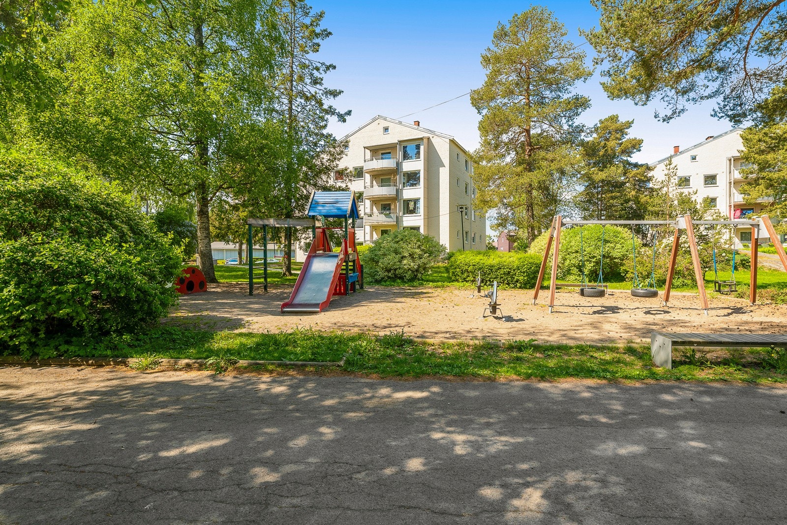 Pent opparbeidet felles tomt med blant annet plener, trær, diverse beplantning, sittebenker med utegrill, lekeplass og asfalterte arealer Galleribilde