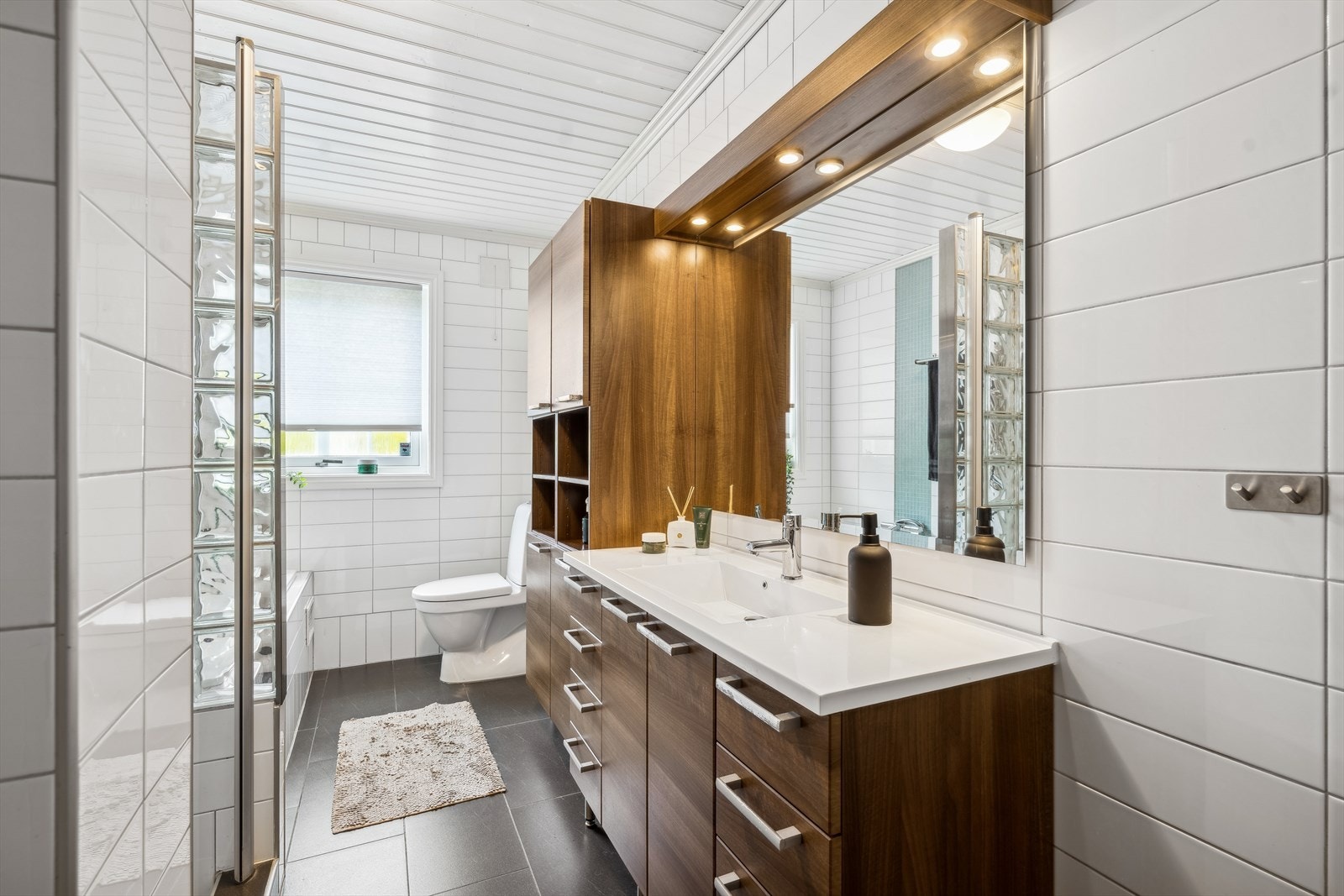 Et velutstyrt og romslig bad med både dusjnisje og innmurt badekar. Dusjnisjen er avskjermet av glassdør og en vegg i glassbyggerstein. Toalett og en bred, helstøpt servant på innredning bestående av underskap, høyskap og speil med belysning. Galleribilde