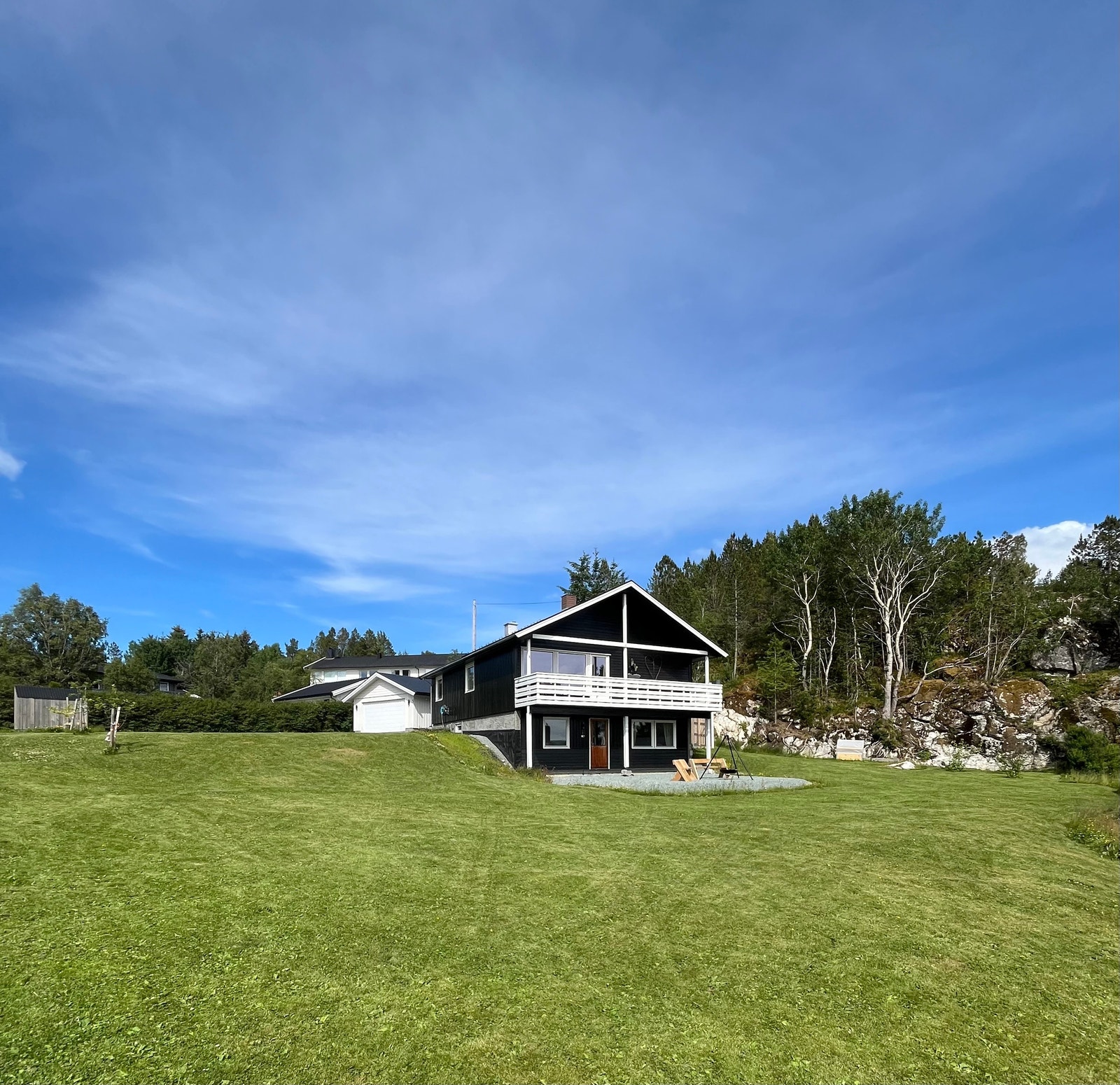 Huset og garasjen sett fra sørvest med plenen foran og uteområdet med benker og bålpanne. Galleribilde