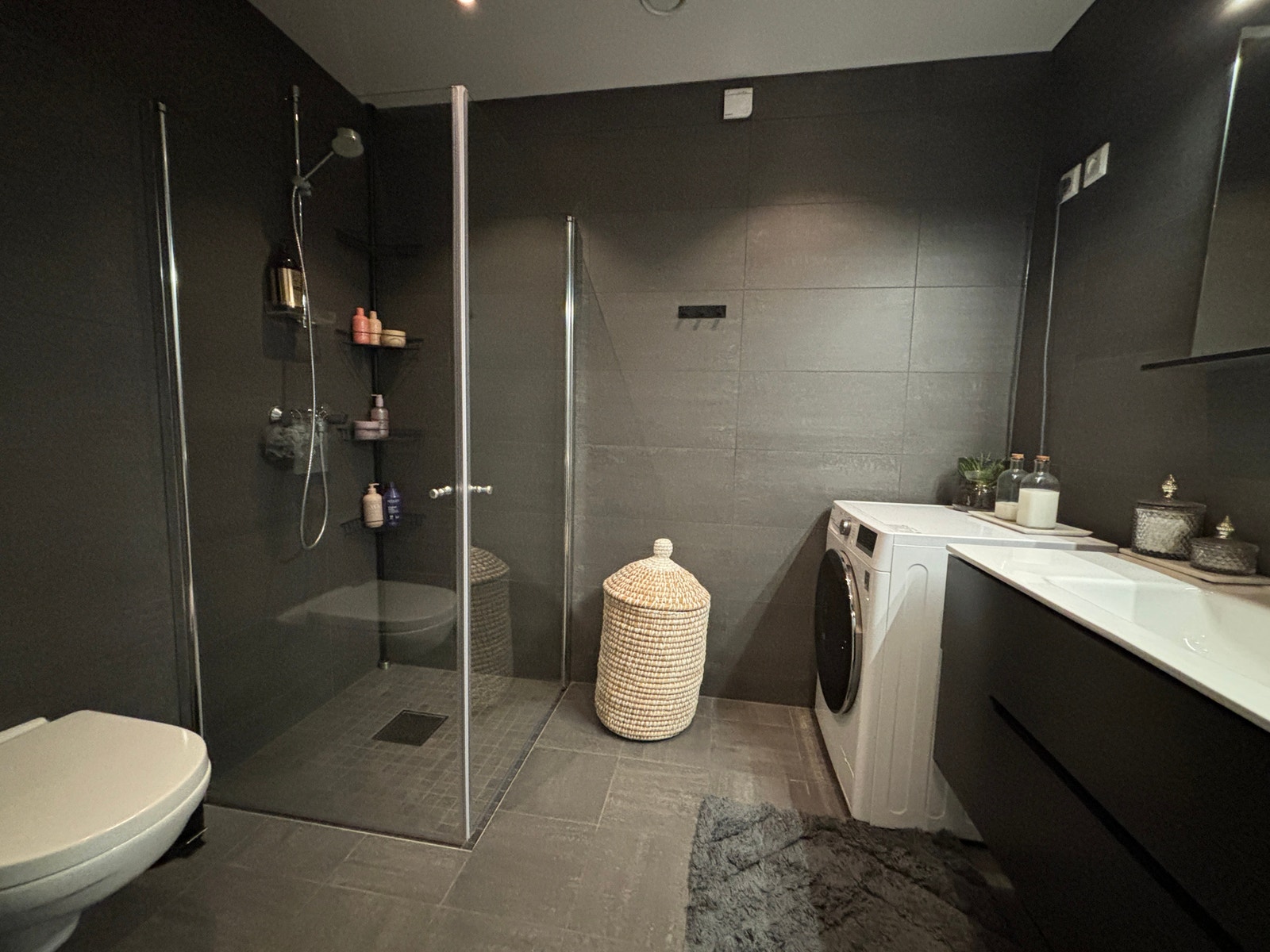 Badet er flislagt vegghengt toalett, dusjhjørne, servant og opplegg for vaskemaskin og tørketrommel. Galleribilde