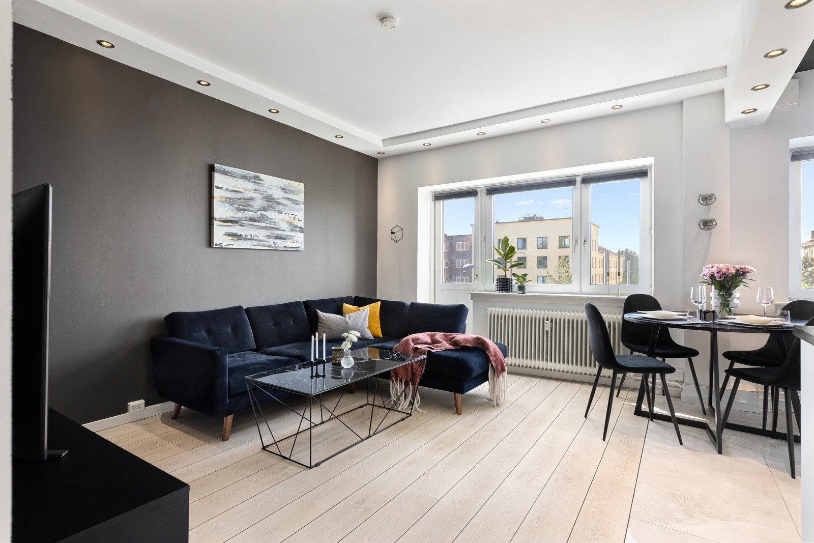 Moderne stue med laminat på gulv, overflater i lyse og mørke fargevalg, og downlights i himlingen. Stuen har flere vindusflater som slipper inn naturlig lys, og fra stuen er det utgang til balkongen. Galleribilde