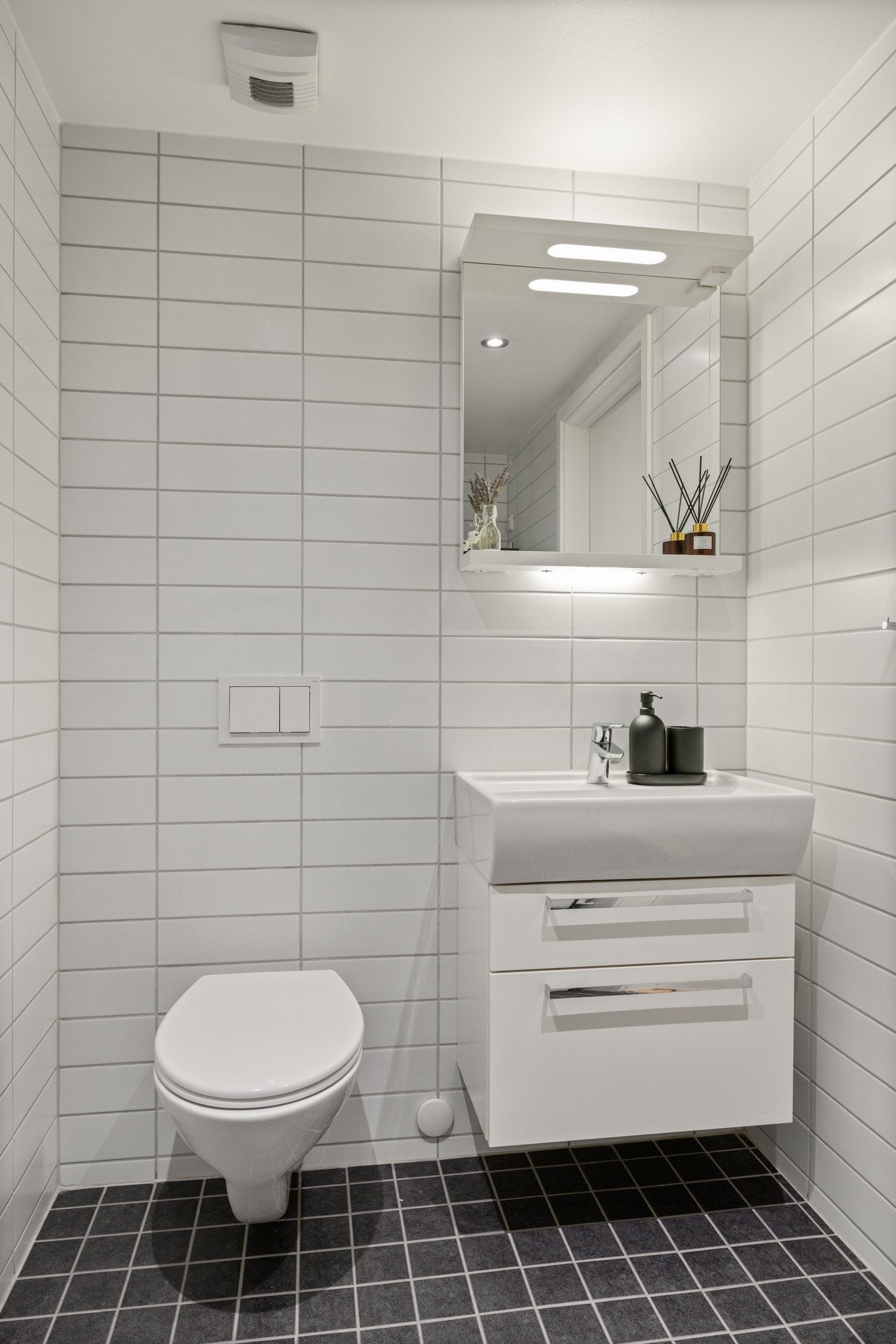 Badet består av nedsenket himling med downlights, vegghengt servantskap med skuffer og ovenpåliggende servant med ett-greps armatur, speil med overlys og stikkontakt på vegg over servant, samt vegghengt toalett. Galleribilde