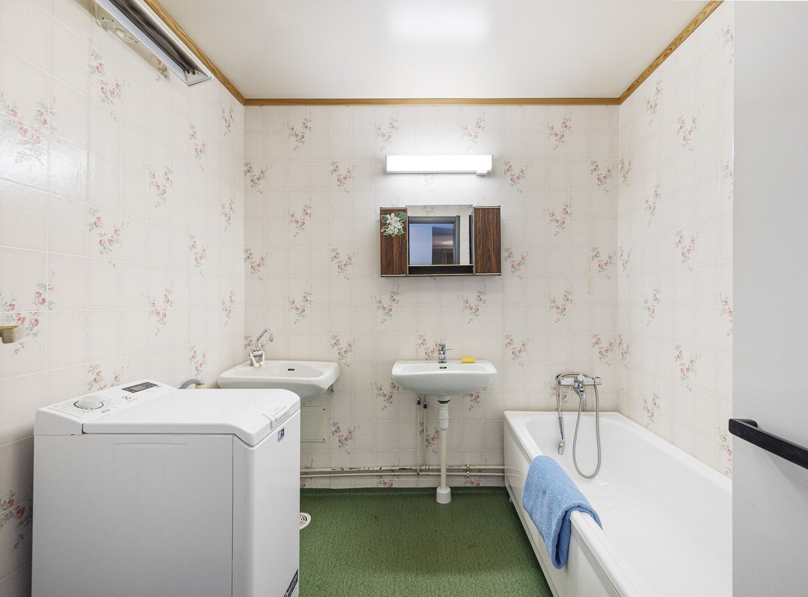 Bad med gulvbelegg på gulv og våtromstapet på vegger. Innredningen består av et høyskap, 2 servanter, speil og armatur med stikk og et badekar. Det er opplegg for vaskemaskin. Galleribilde