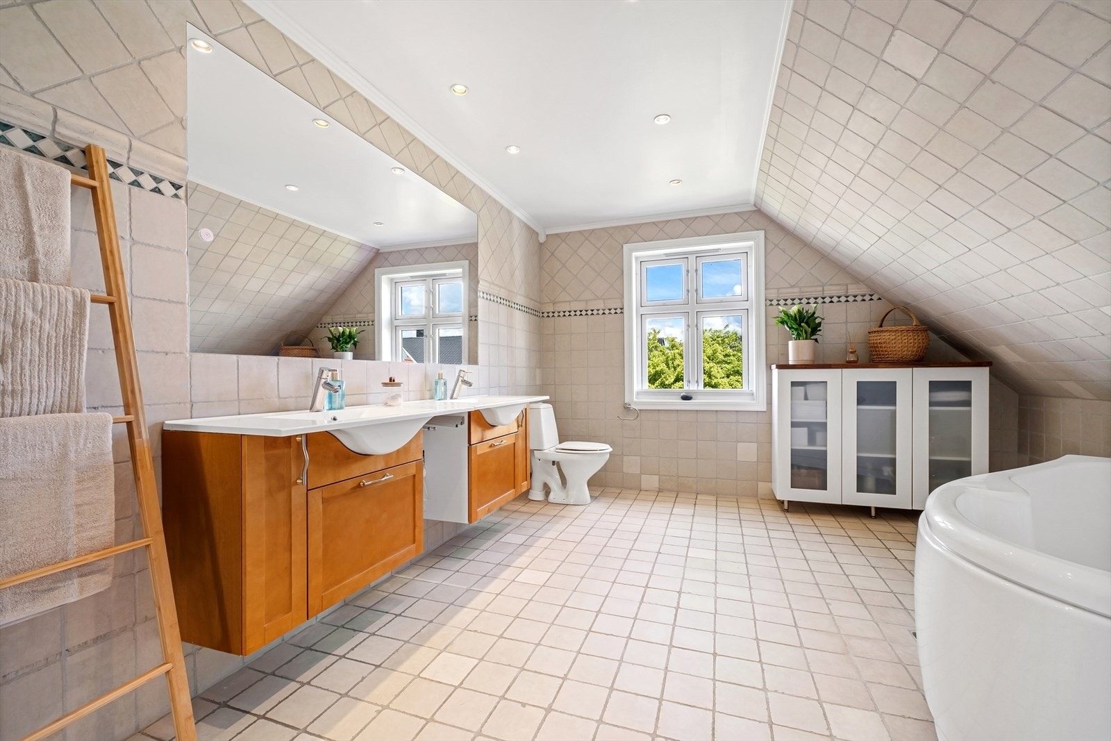 Romslig hovedbad som har innredning med helstøpt benk med to servanter, stort innfelt speil og gulvmontert toalett Galleribilde