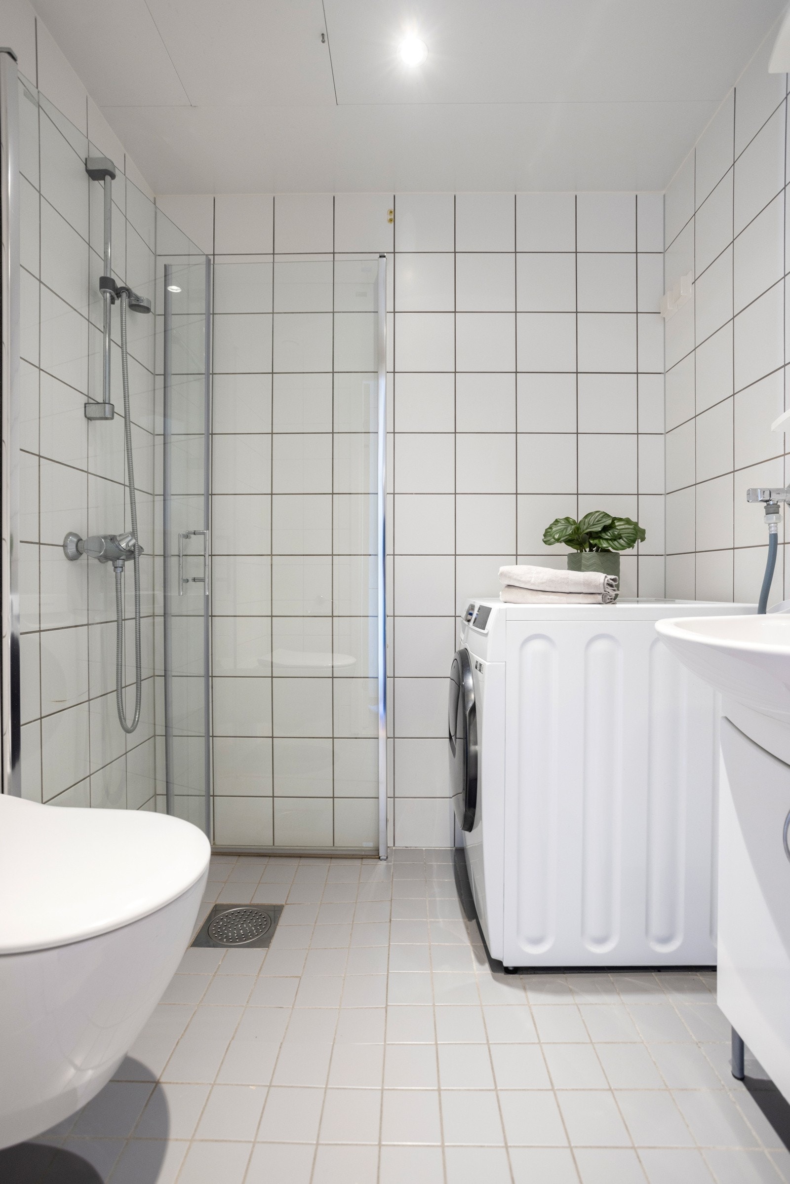 Lyst flislagt bad med varmekabler i gulv. Speilskap med lysarmatur, vegghengt klosett, dusjhjørne med svingdører i glass og opplegg for vaskemaskin Galleribilde