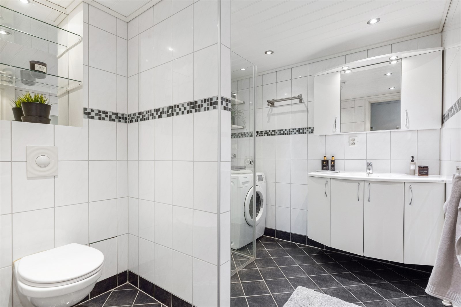 Badet har et vegghengt toalett, opplegg for vaskemaskin og et dusjhjørne med innfellbare glassdører fra 2017. Galleribilde