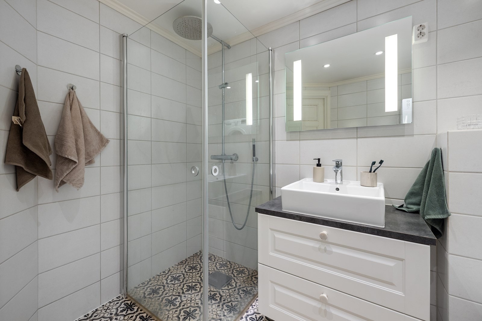 Badet har innredning og garnityr som består av baderomsinnredning med skuffer, servant, ett-greps blandebatteri fra Hansgrohe og speil, veggmontert toalett og dusjhjørne med innfellbare dører Galleribilde