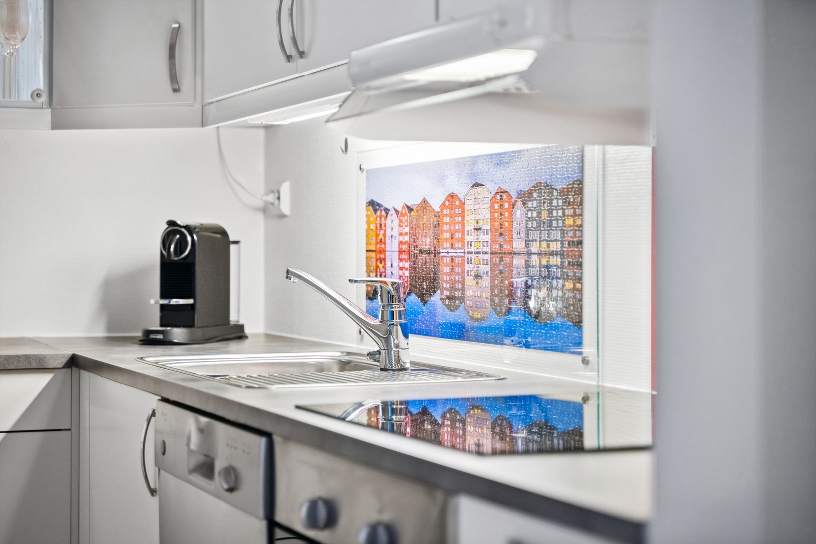 Kjøkkenet er innredet med hvite slette kjøkkenfronter, laminat bankeplate, nedfelt vask med benkeslag, integrert stekeovn og platetopp. Frittstående oppvaskmaskin og kjøleskap. Pleksiglassplate over vask og platetopp med motiv fra Trondheim. Galleribilde
