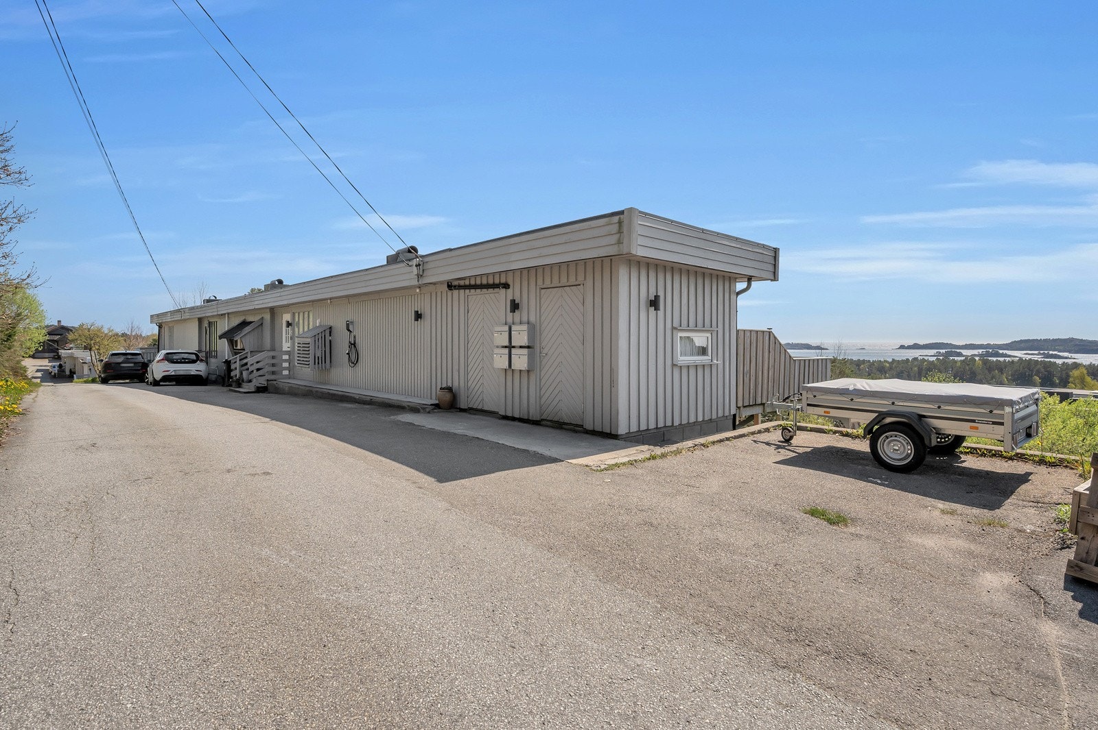 Fasade sett fra veien med biloppstillingsplasser langs husveggen og utstikker. Halve bod ytterst til høyre tilhører nabo. Galleribilde