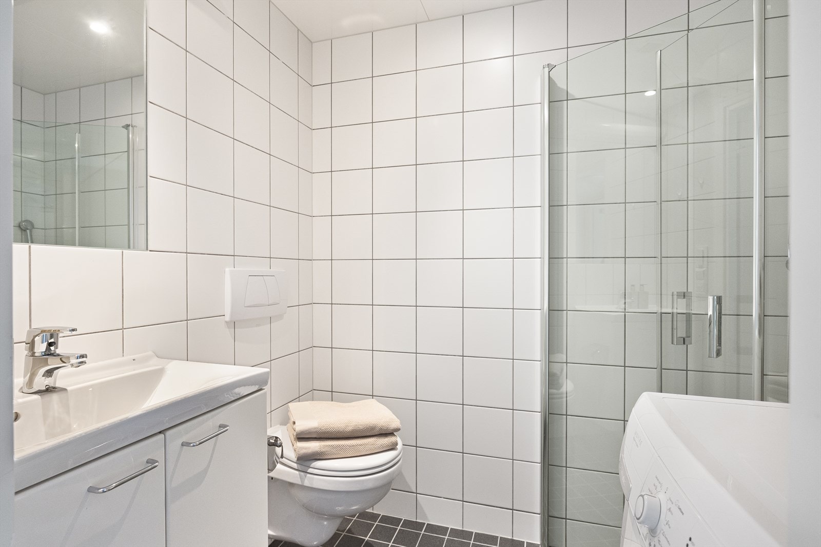 Badet har en funksjonell innredning, inkludert en flott servant med skuffeinnredning og et speil med integrert belysning over. Videre er det vegghengt toalett og en dusjnisje med innfellbare glassdører, som gir en romslig og praktisk dusjløsning. Galleribilde