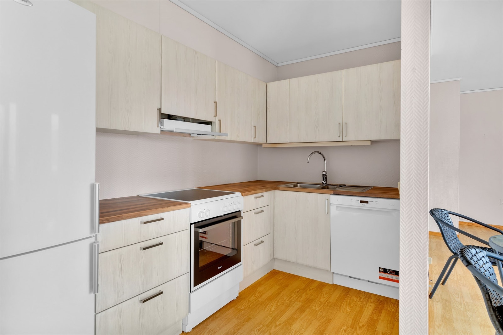 Åpen og sosial løsning mellom stue og kjøkken. Hvitevarene består av frittstående kjøleskap med frysedel, komfyr og oppvaskmaskin under benkeplate. Det er avtrekksvifte over komfyr med mekanisk avtrekk. Galleribilde