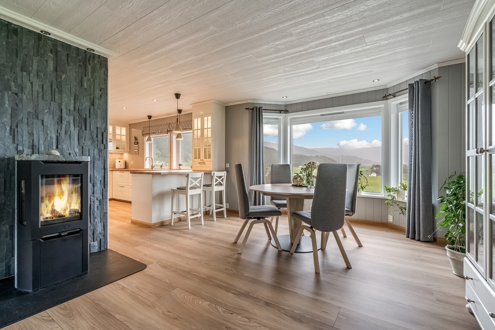 Stue med klebersteins vedovn med glass på tre sider og fin brannmur i pynteskifer. Plissè gardiner i vindu og spotter i tak. Galleribilde