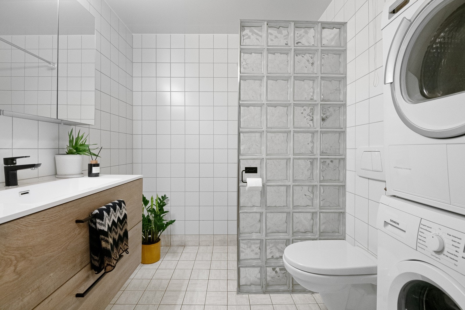 Badet er flislagt med varmekabler i gulv, veggmontert toalett og dusjhjørne med glassbyggerstein. Opplegg til vaskemaskin og plass for tørketrommel i en praktisk nisje som gir maksimalt utbytte av plassen. Galleribilde