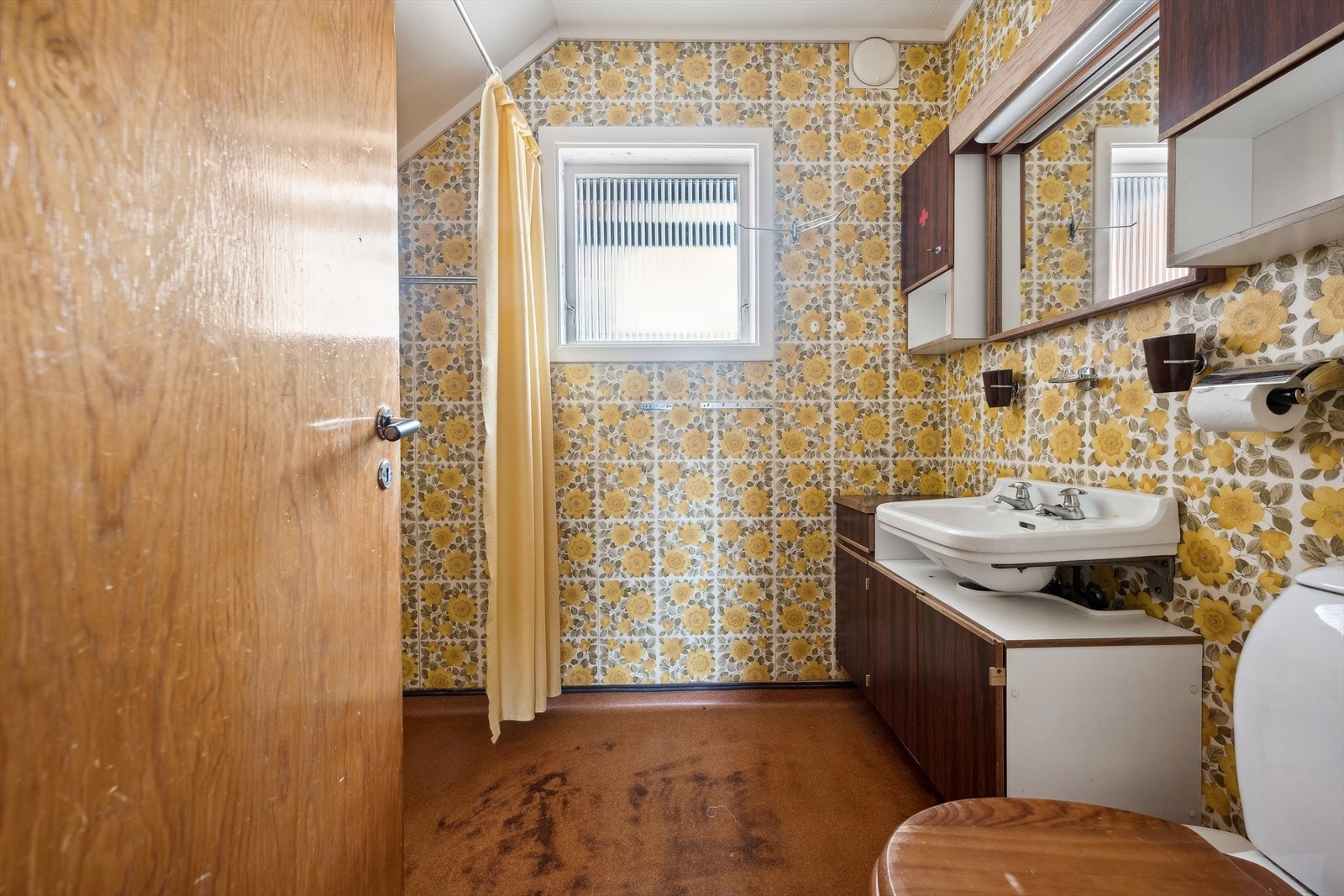 Badet har tapet på veggene og malte plater/takess i taket. På gulvet er det belegg. Rommet har et vegghengt baderomsmøbel med vask, frittstående toalett og dusjhjørne. Galleribilde