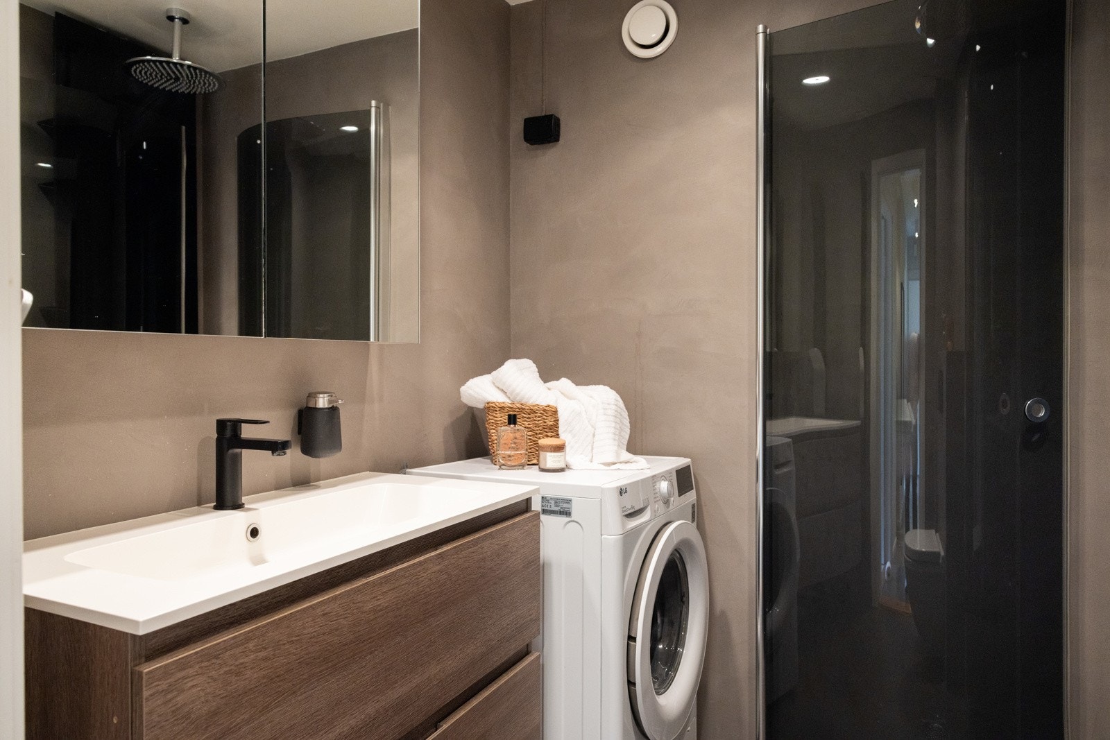 Rommet er innredet med gulvmontert toalett, nedsenket vask med skuffer og speil, en dusjnisje med glassdører, og opplegg for vaskemaskin. Galleribilde