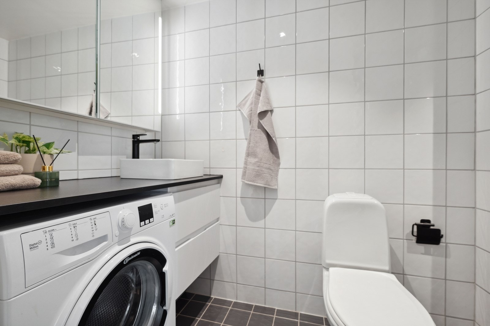 Pent fliselagt baderom bestående av frittstående toalett, servant med underskap, overskap med speilfronter og lys, inn-/utadslående dusjdører og opplegg for vaskemaskin Galleribilde