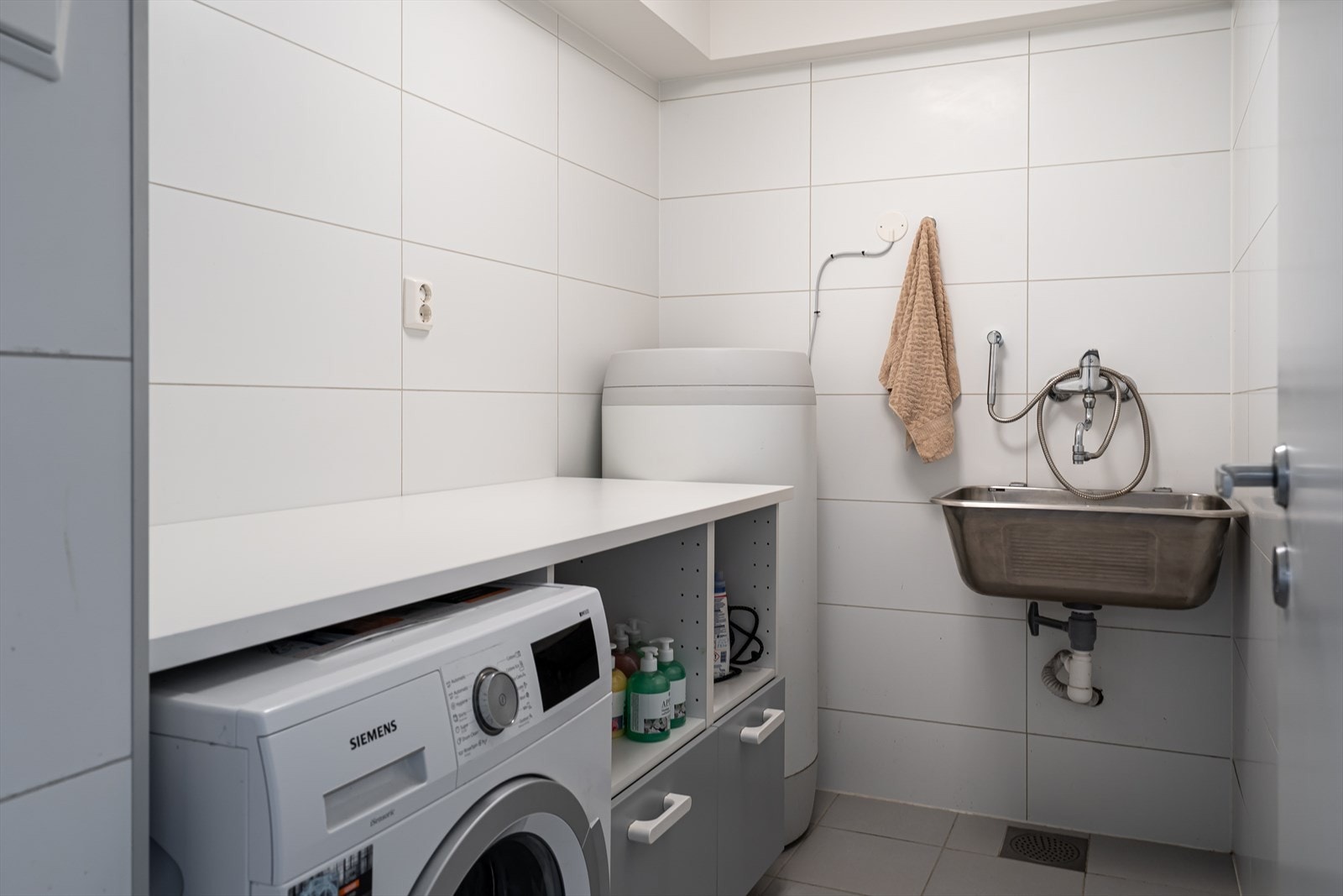 Vaskerommet ligger vegg-i-vegg med badet og har grå gulvfliser, hvite veggfliser og gulvvarme. Galleribilde