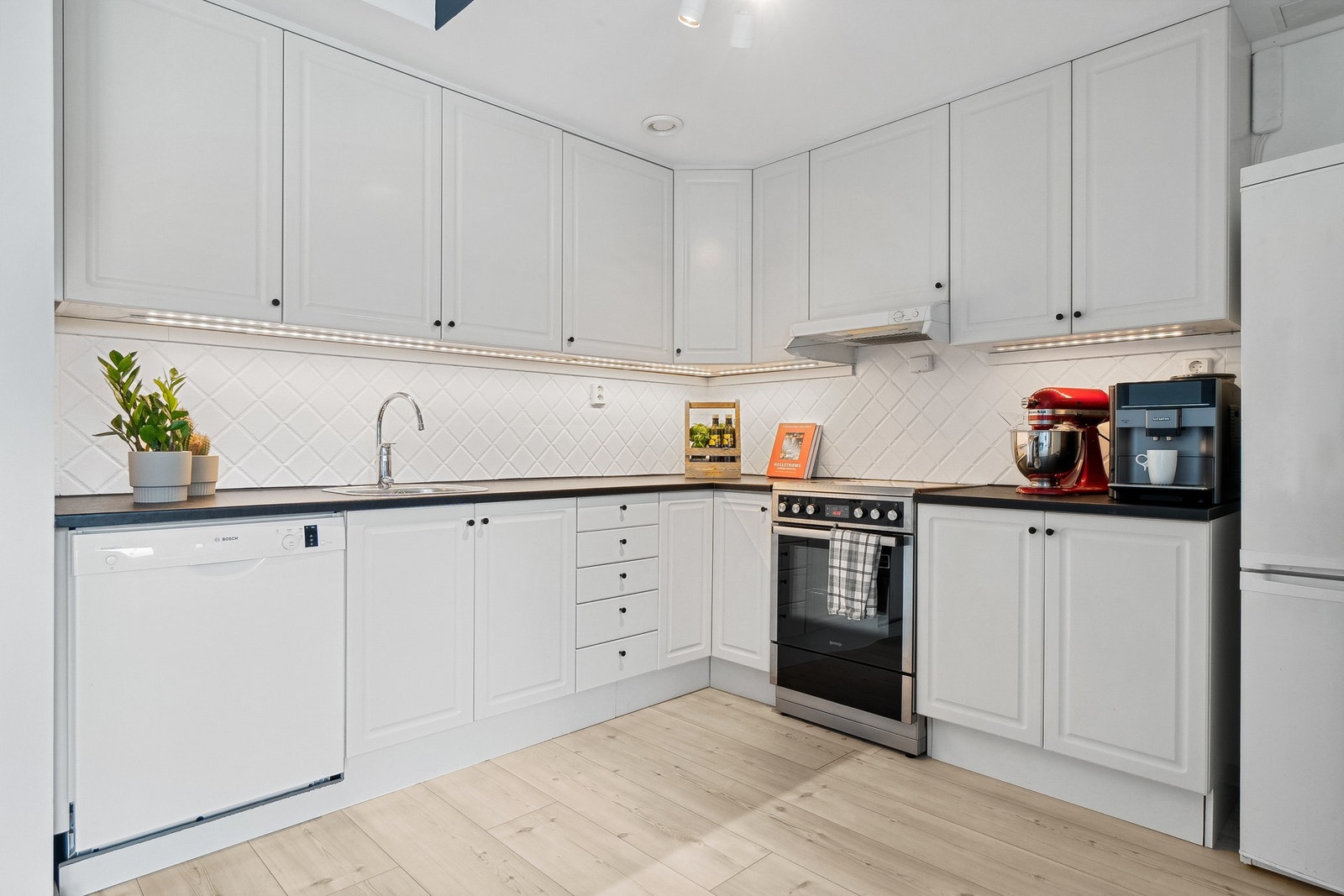 Lys og pen kjøkkeninnredning med takhøye skap, flis over benk og opplegg for oppvaskmaskin. Galleribilde